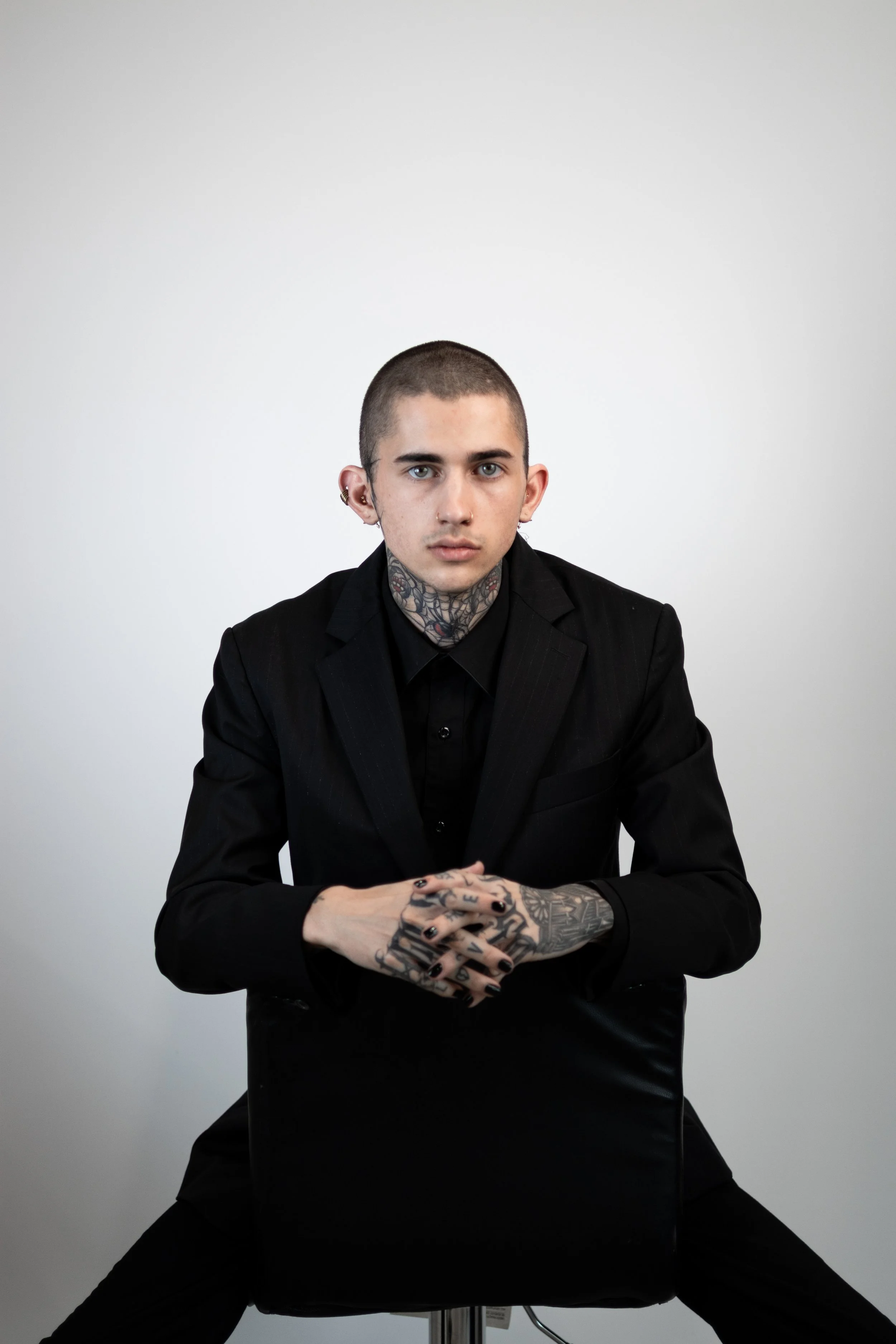 A young man with tattoos on his neck and hands, wearing a black suit and seated on a black chair, against a plain gray background. Specializes In Alternative Women Haircuts and Vivid Hair Color.