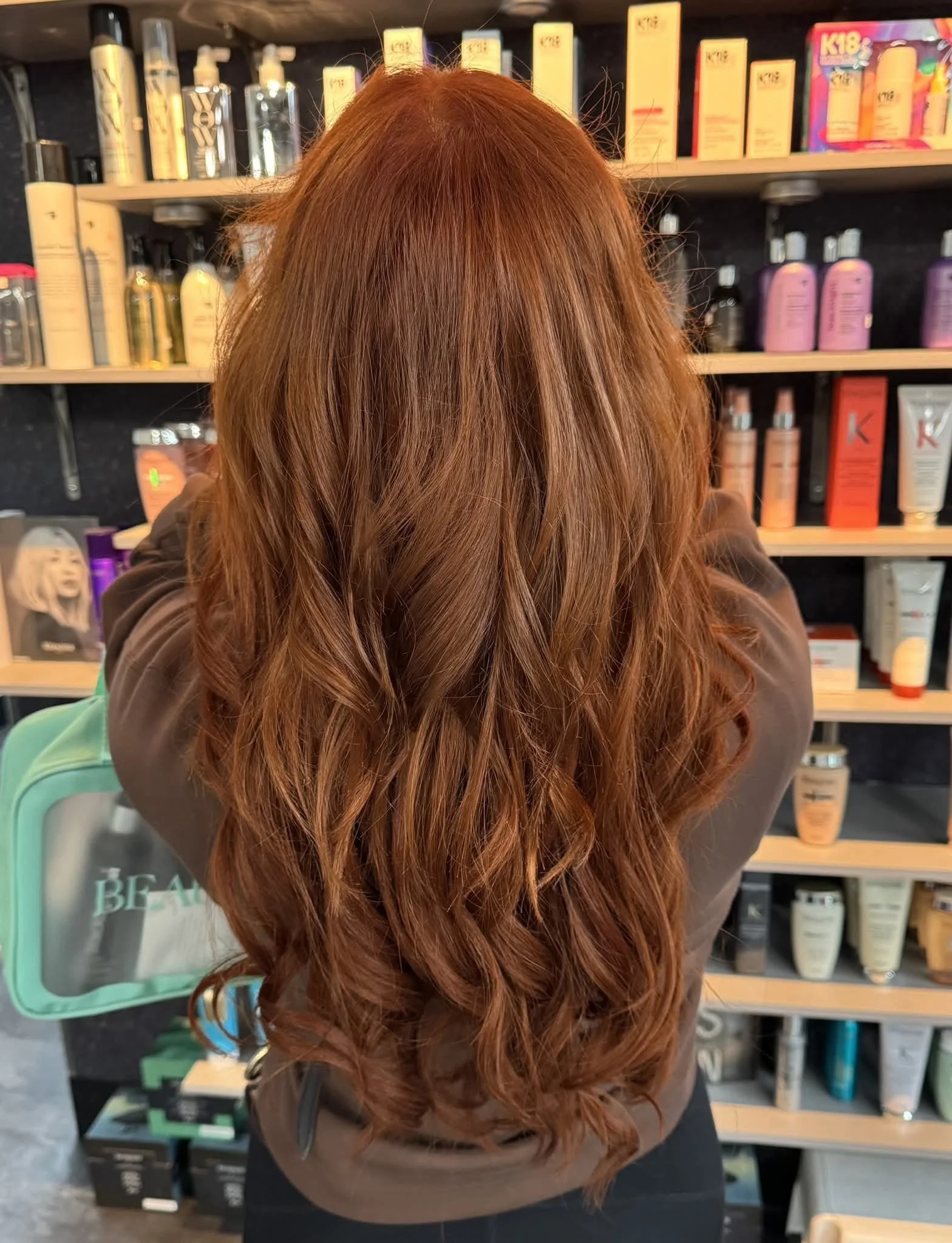 Person with long, wavy, reddish-brown hair standing in front of shelves stocked with hair care and beauty products .Long layered hair extensions with natural blending at Misfits Salon