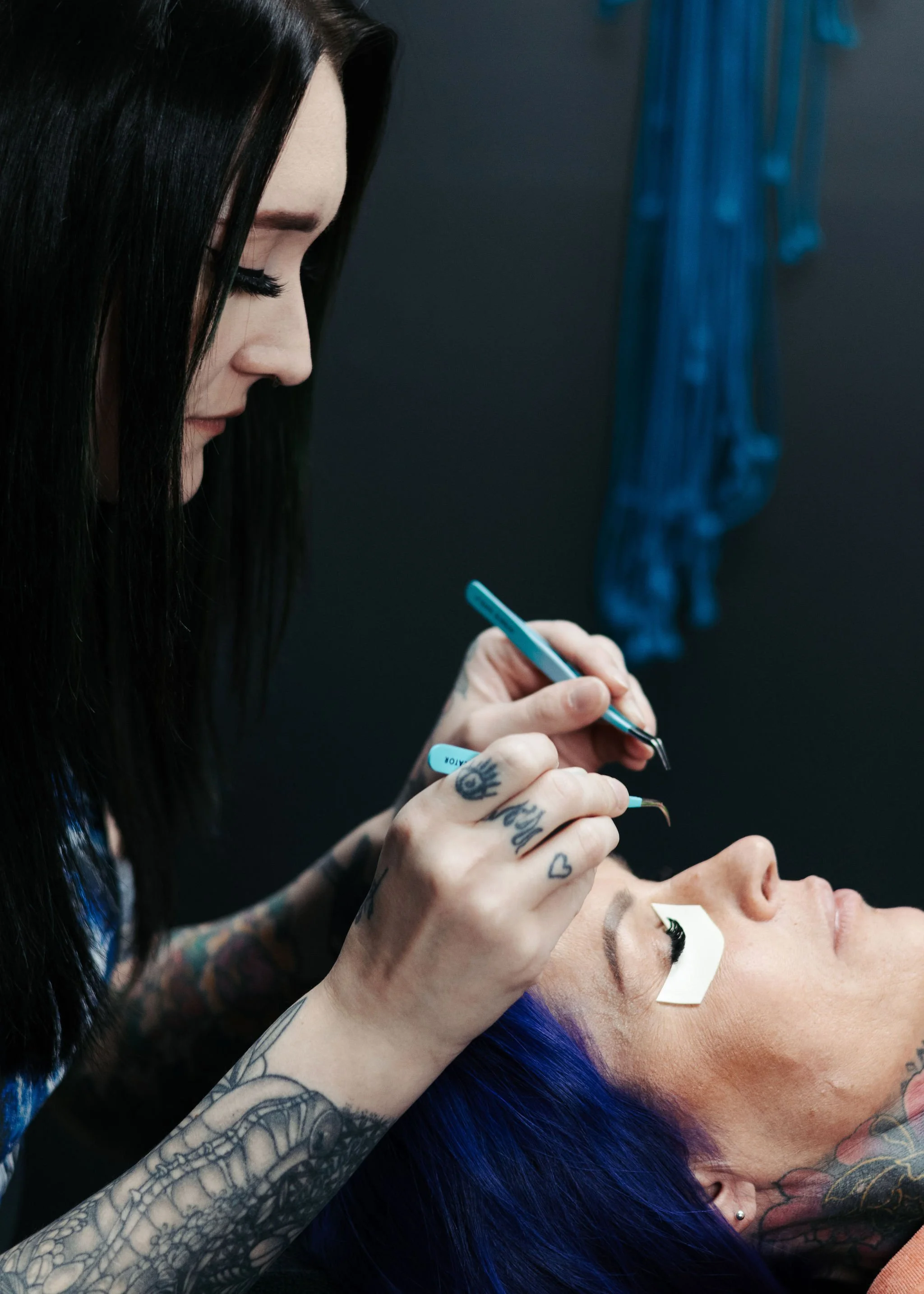 A tattooed woman with blue hair getting eyelash extensions from a technician with tattoos on her hands. The woman is lying back with eyes closed, and getting hybrid lash extensions at Misfits Salon in Anchorage Alaska. 