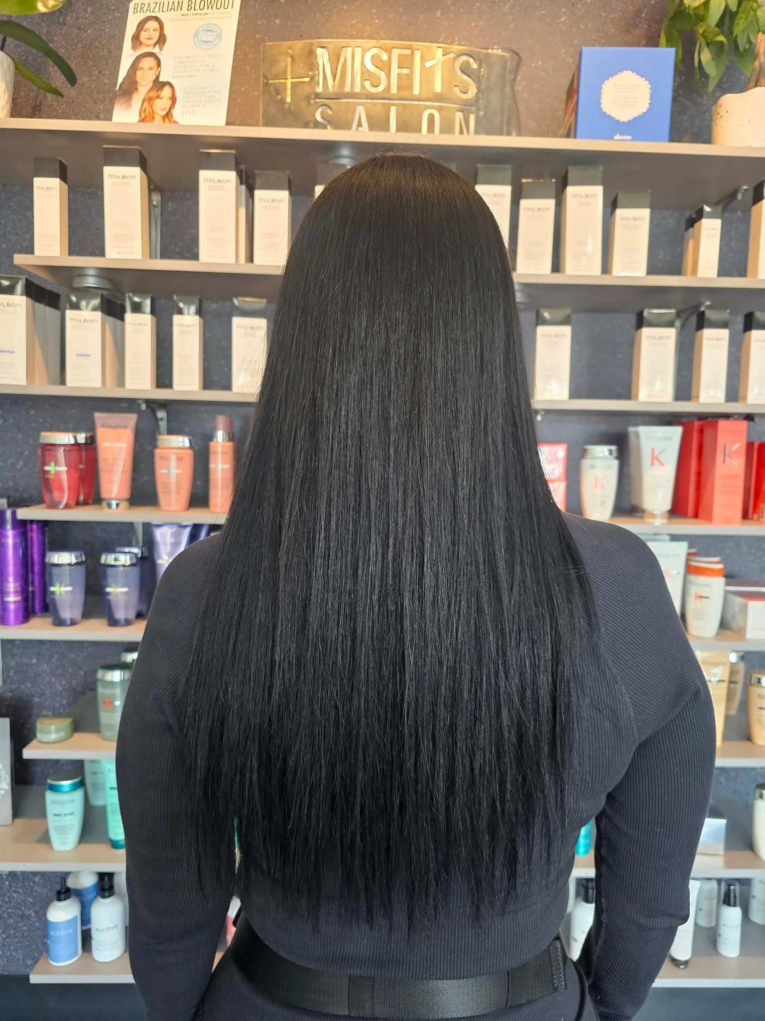 The image shows a woman with long, straight black hair extensions wearing a black top, standing in front of shelves filled with hair and skincare products in a salon. The salon sign is visible in the background.