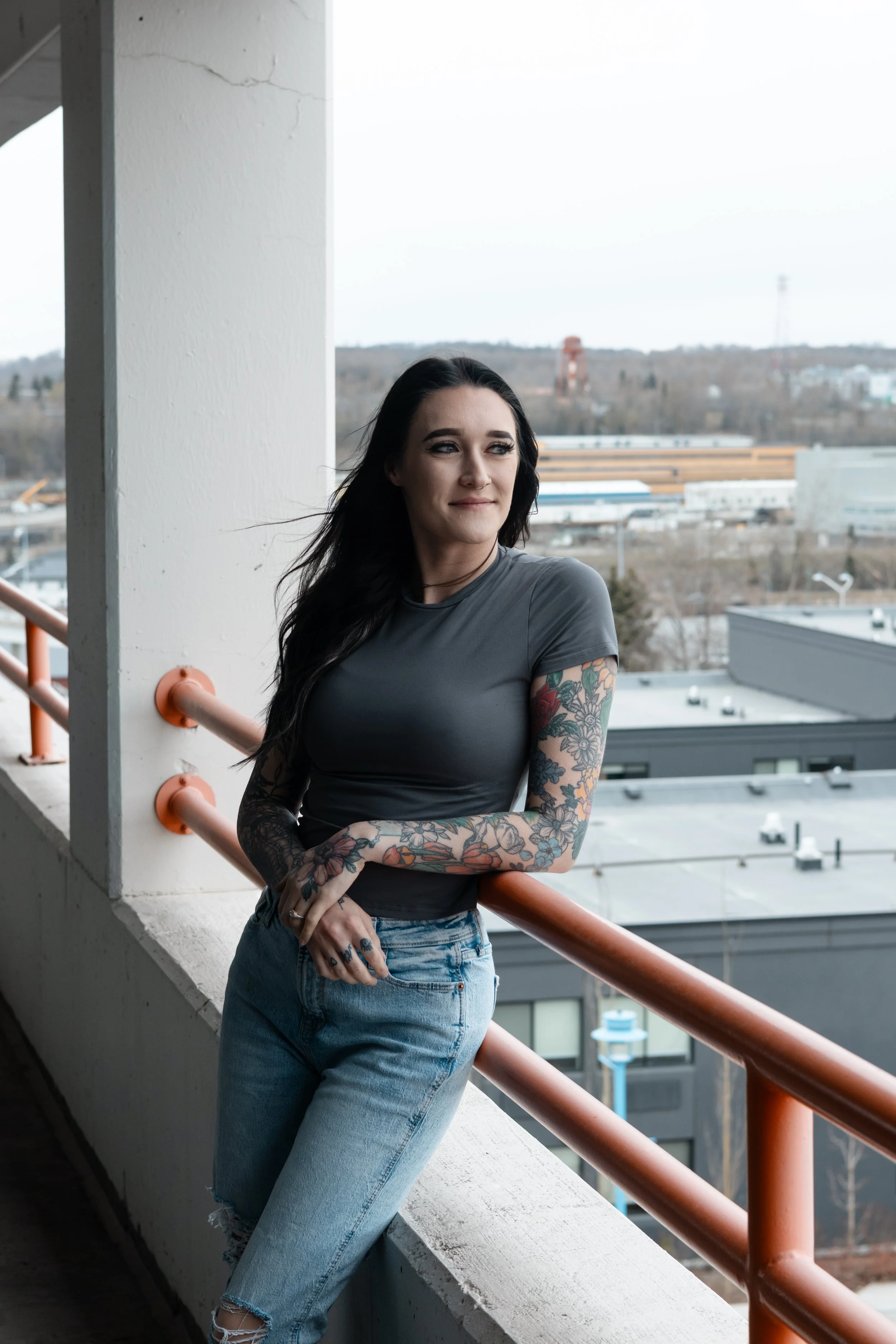 A young woman with long dark hair, tattoos on her arms, wearing a dark gray T-shirt and light blue jeans, standing on a balcony with a city view behind her. She is a Lash Artist at Misfits Salon and specializes in waxing and lash services. 