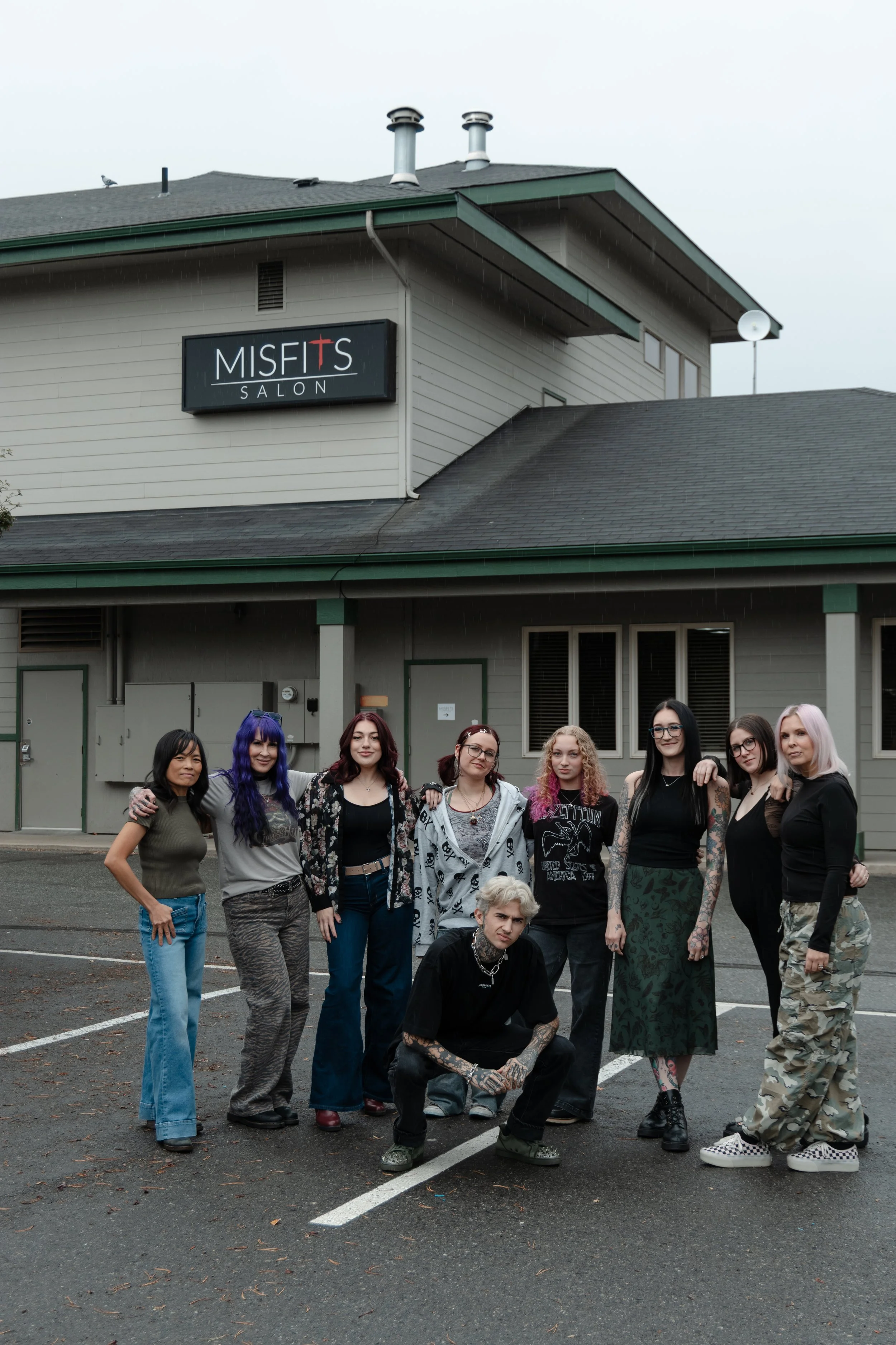 Group of diverse young people posing outdoors in front of a building with a sign that reads 'Misfits Salon' on a cloudy day. The professional beauty team at Misfits Salon in Anchorage, Alaska, offering luxury hair extensions, balayage, $ color.