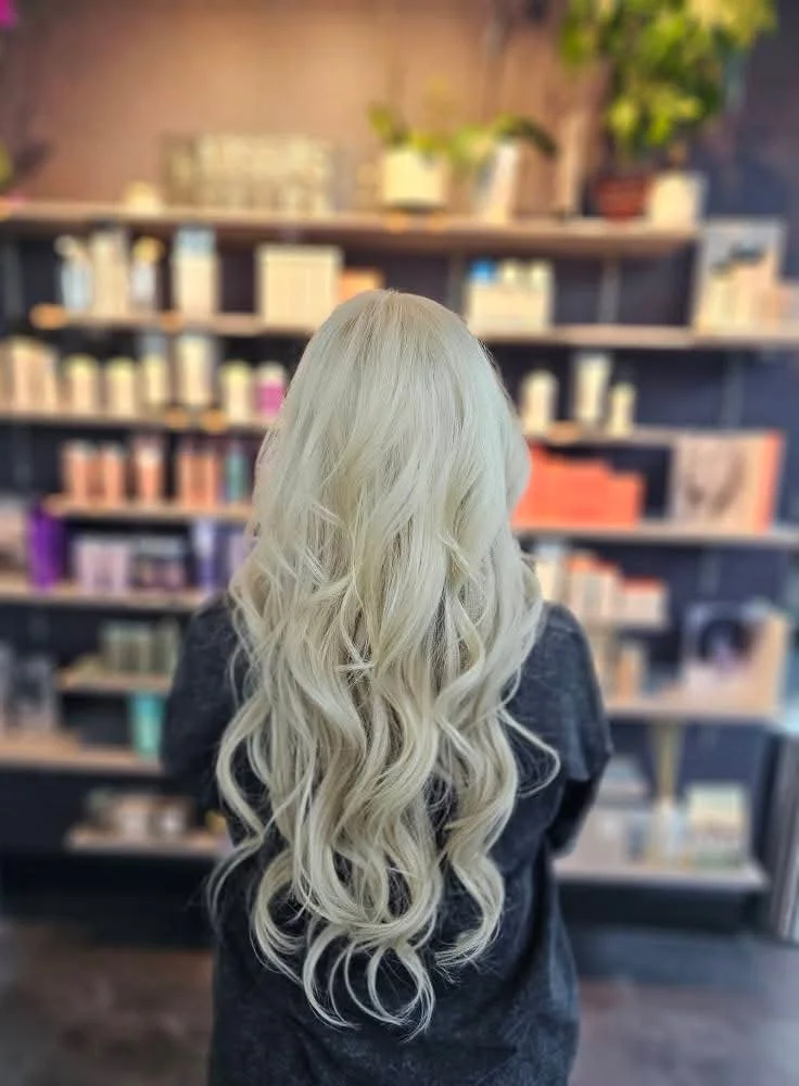 Person with long, wavy, platinum blonde hair extensions standing in front of a blurred bookshelf.
