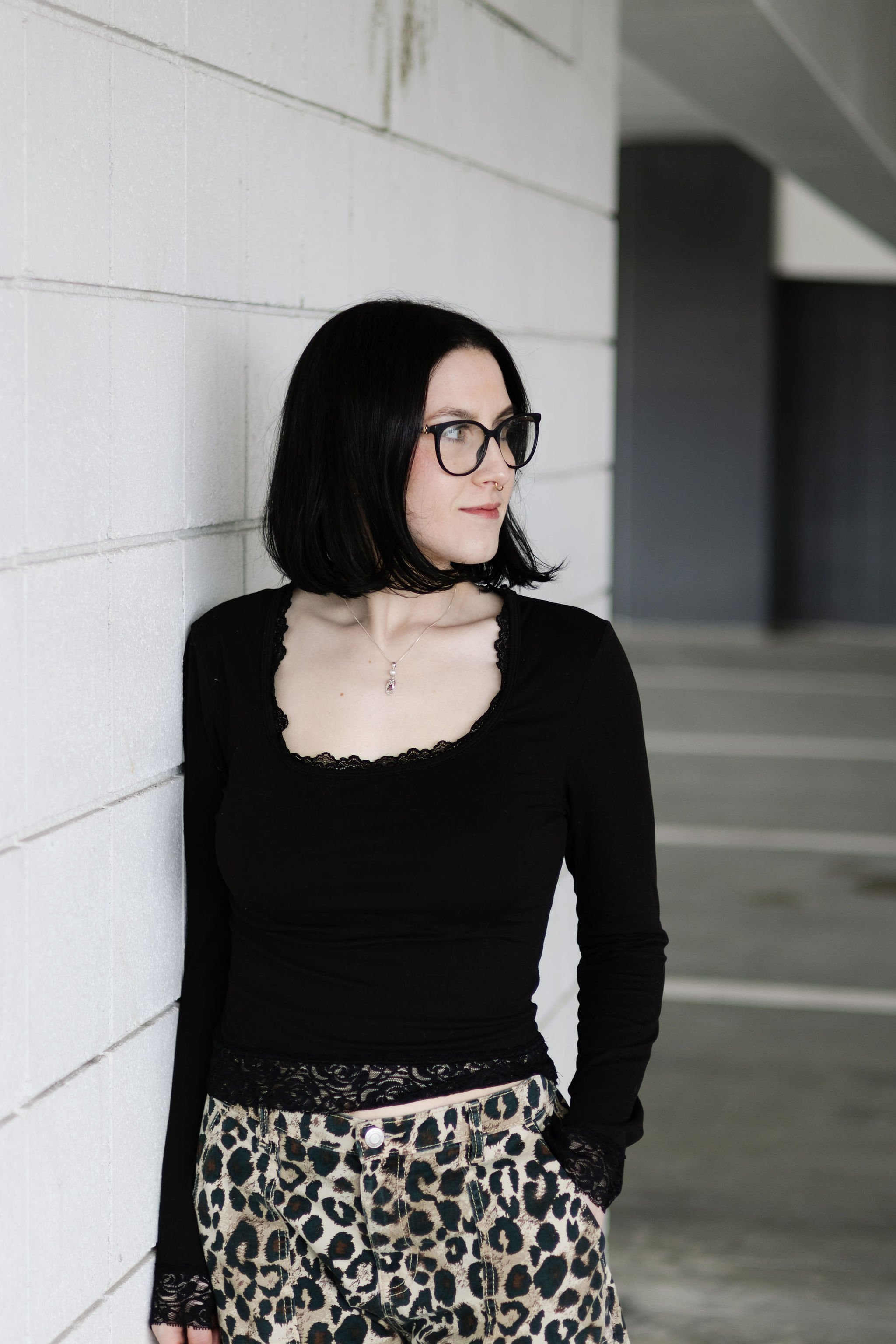 A woman with black hair and glasses standing against a white cinder block wall, looking to the right, wearing a black top and animal print skirt. She specializes in Alternative Hair Styles and vivid color.She offers a range of salon services includin