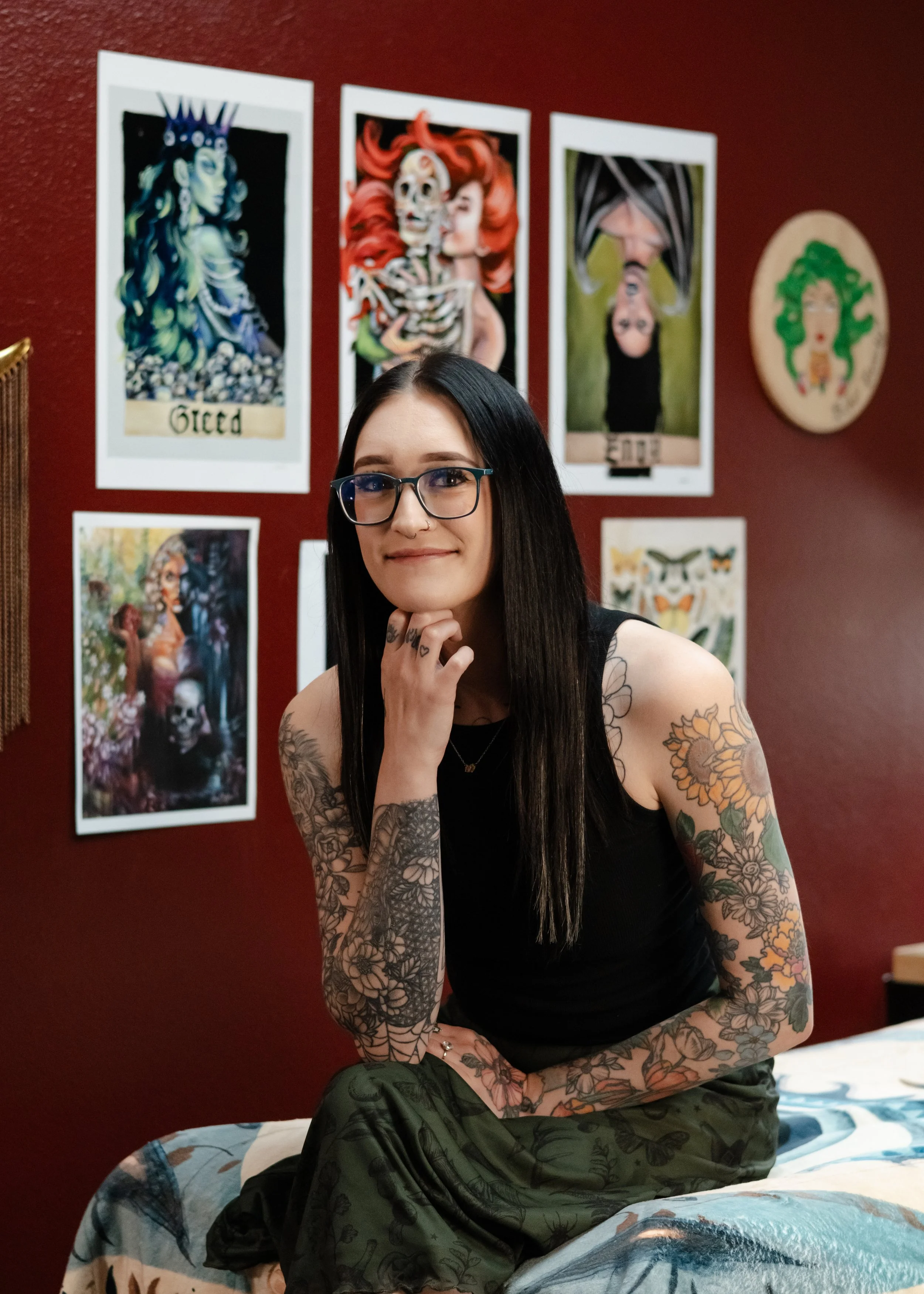 A young woman with long black hair and glasses, smiling and resting her chin on her hand. Hunter, master esthetician at Misfits Salon in Anchorage, Alaska, specializing in lash extensions, lash lifts, Brazilian waxing, & professional waxing services.