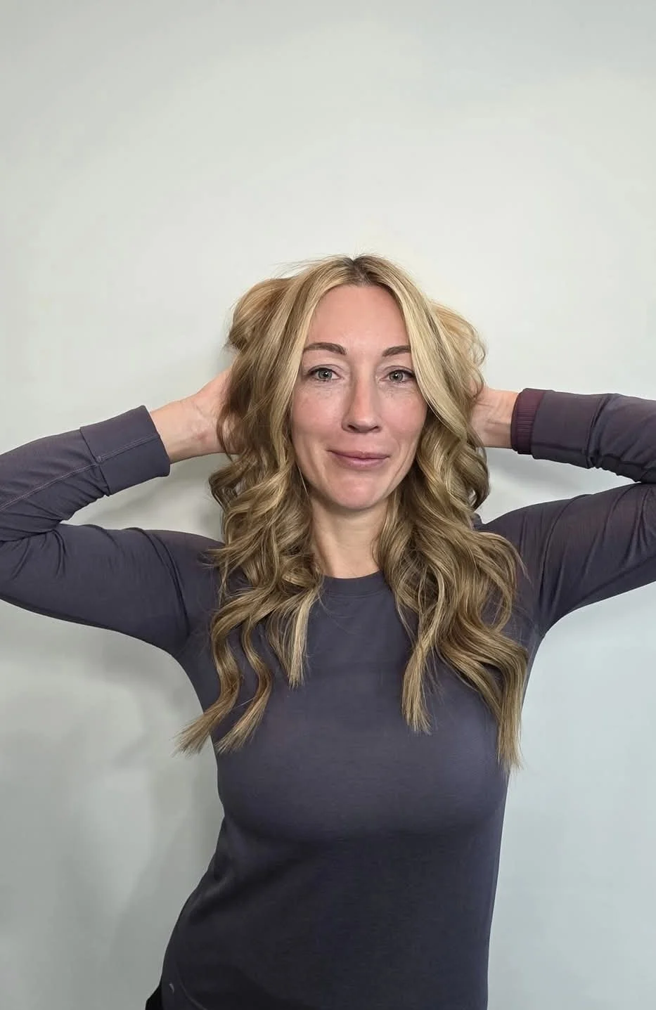 Woman with long, wavy blonde hair, wearing a dark long-sleeve shirt, smiling and holding her hair back with her hands.