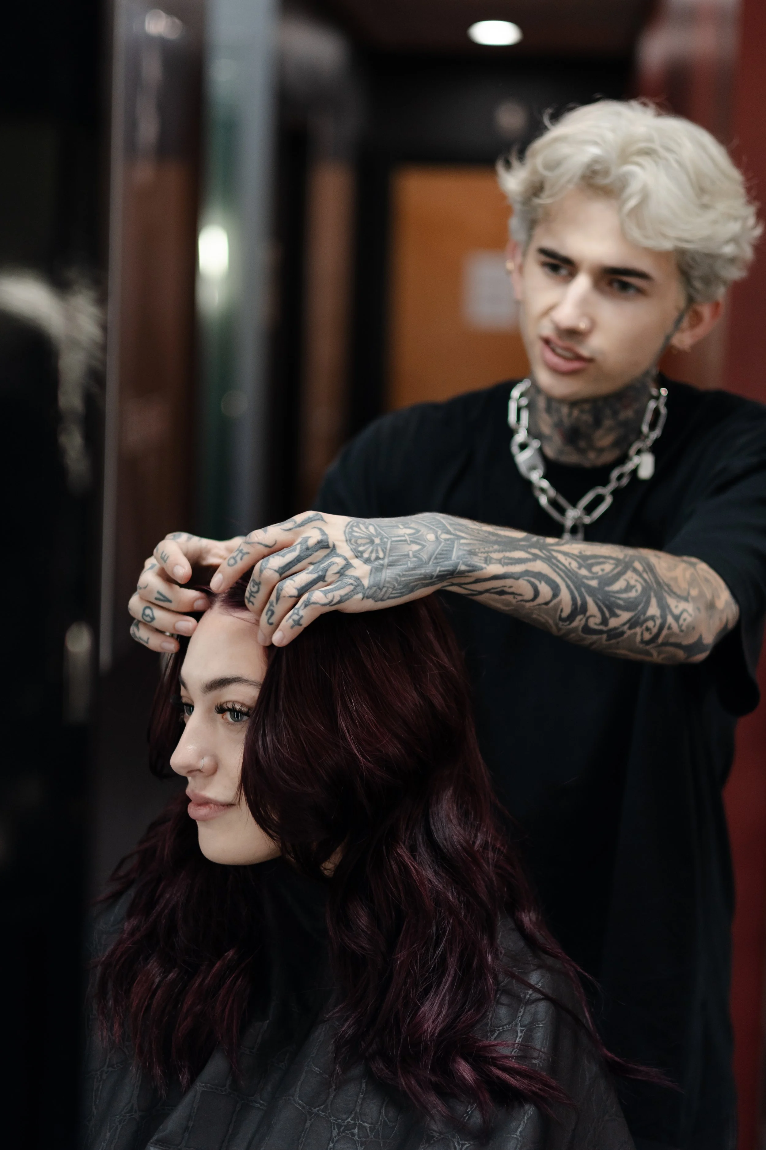 A hairstylist with tattoos and bleached hair styles a woman with dark red hair in a salon. Professional hairstylists at Misfits Salon in Anchorage, Alaska, offering luxury hair extensions, custom hair color, balayage, blonding & vivid colors.