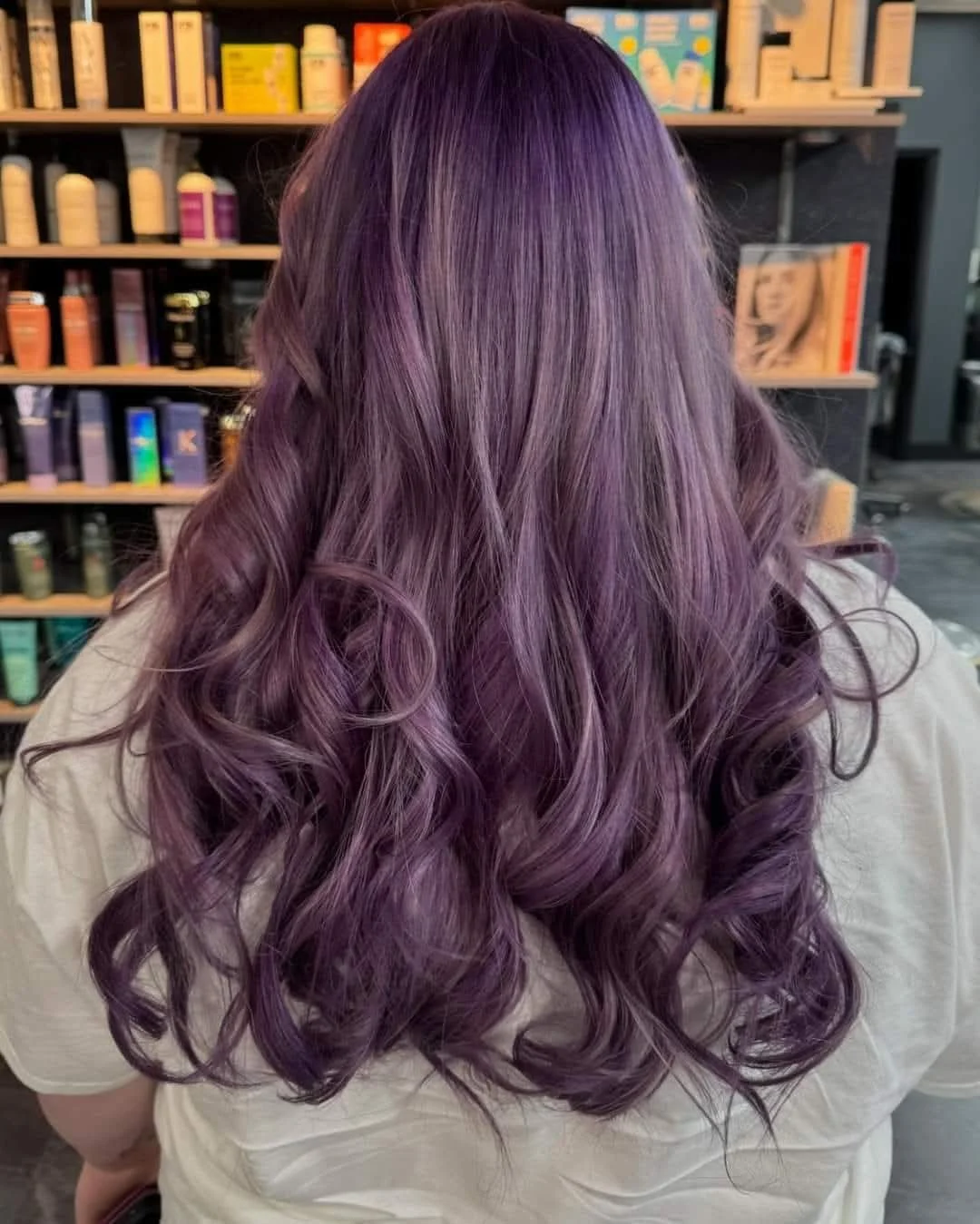 Back view of a person with long, wavy hair colored in shades of purple and lavender, in a salon or store with shelves of hair care products in the background.