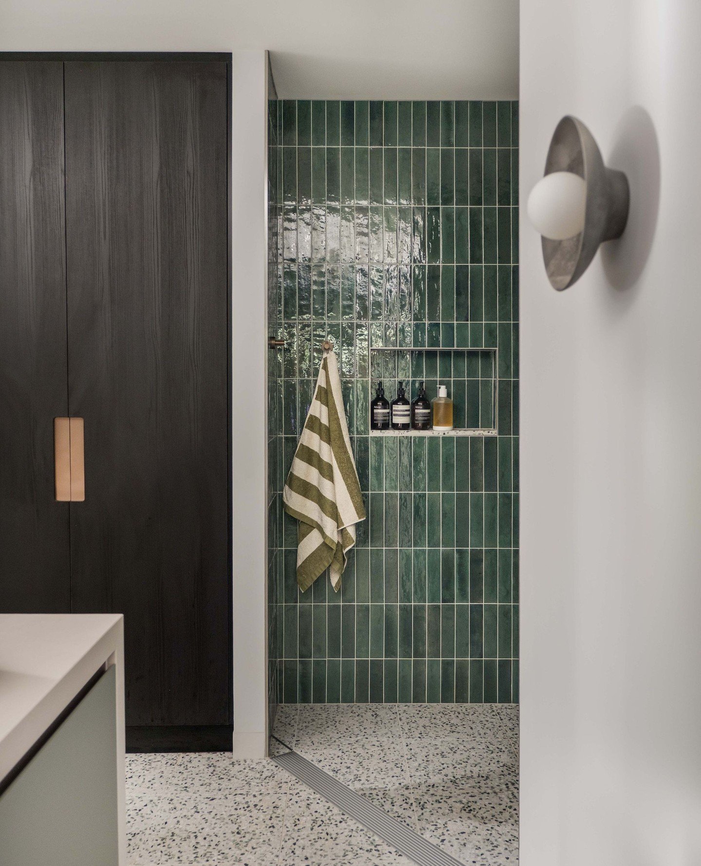 GREAT ALPINE ROAD⁠. This parents' retreat was inspired by a hotel suite. A curved, green-tiled shower becomes a sculptural centrepiece, offset by terrazzo flooring and dark timber joinery with brushed-brass accents. Floating mirrors above a custom is