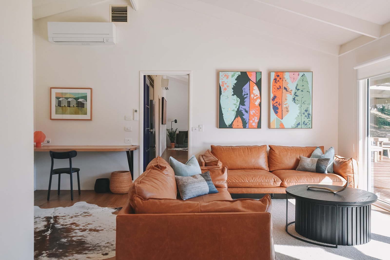 Inviting holiday home living room featuring warm tan leather sofas, colourful botanical artwork, contemporary furniture and a dedicated workspace.