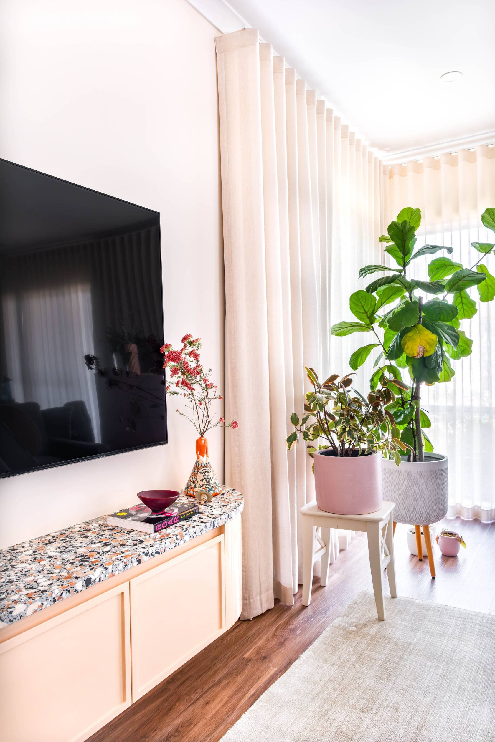 The custom entertainment unit continues the terrazzo story in the living room, layered with soft pastel sheers, warm flooring, plants and light.