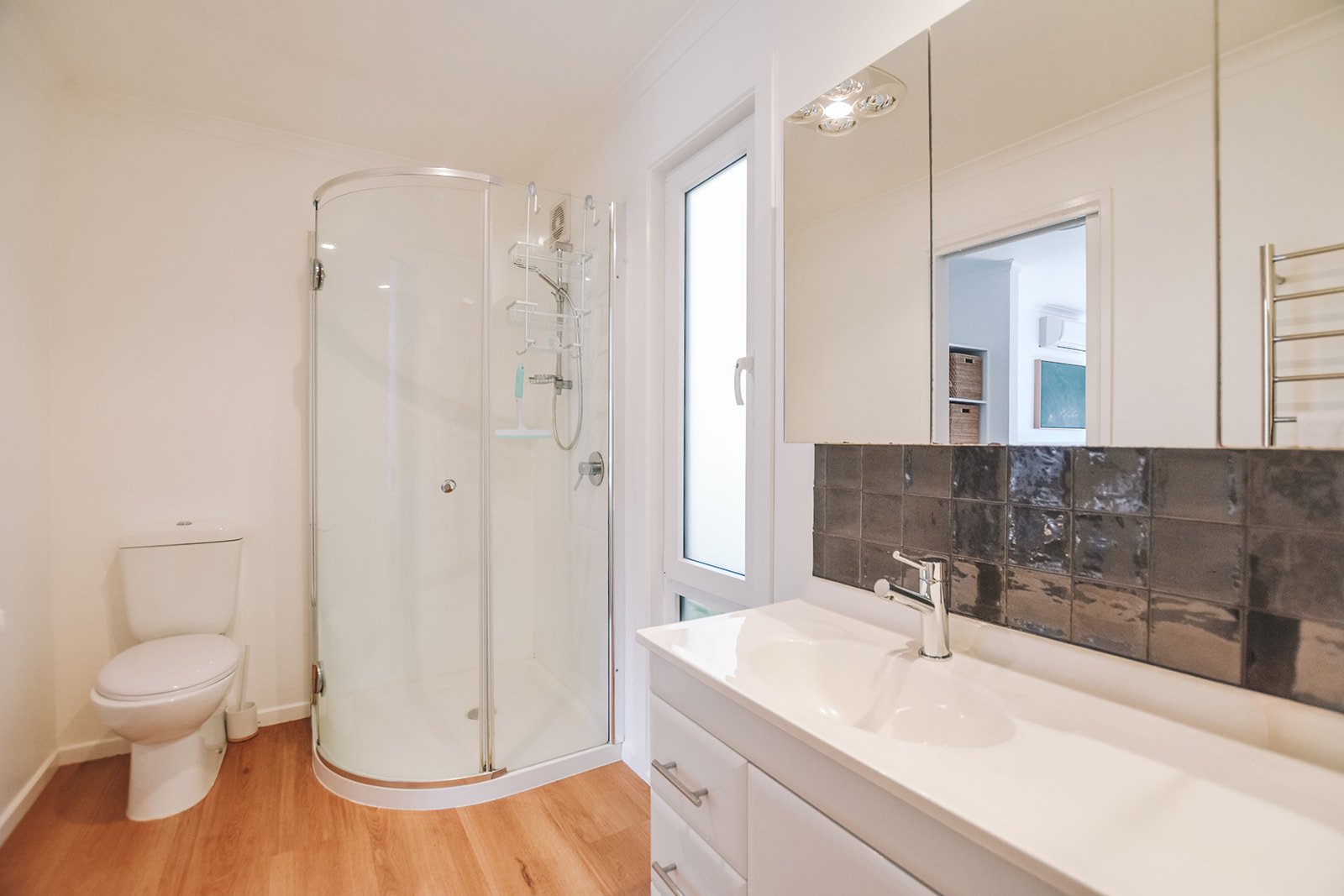 Bright holiday home bathroom featuring white cabinetry, textured dark splashback tiles, contemporary tapware and timber flooring.
