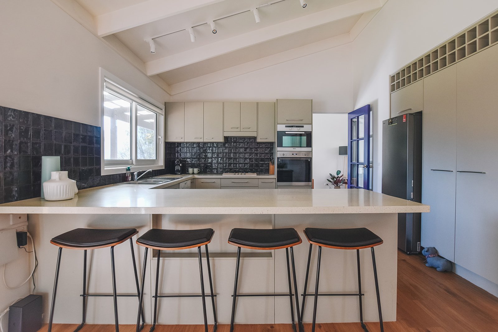 Refreshed holiday home kitchen featuring textured black splashback tiles, neutral cabinetry, stone benchtops and contemporary black bar stools.