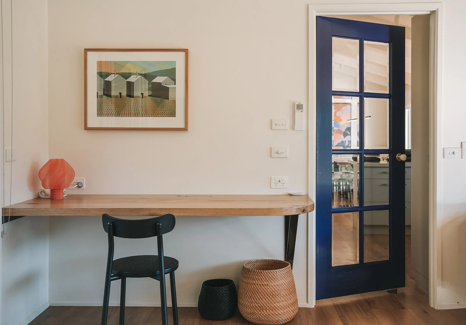 Compact home workspace featuring a natural timber desk, black chair, colourful artwork and a bold blue internal door.