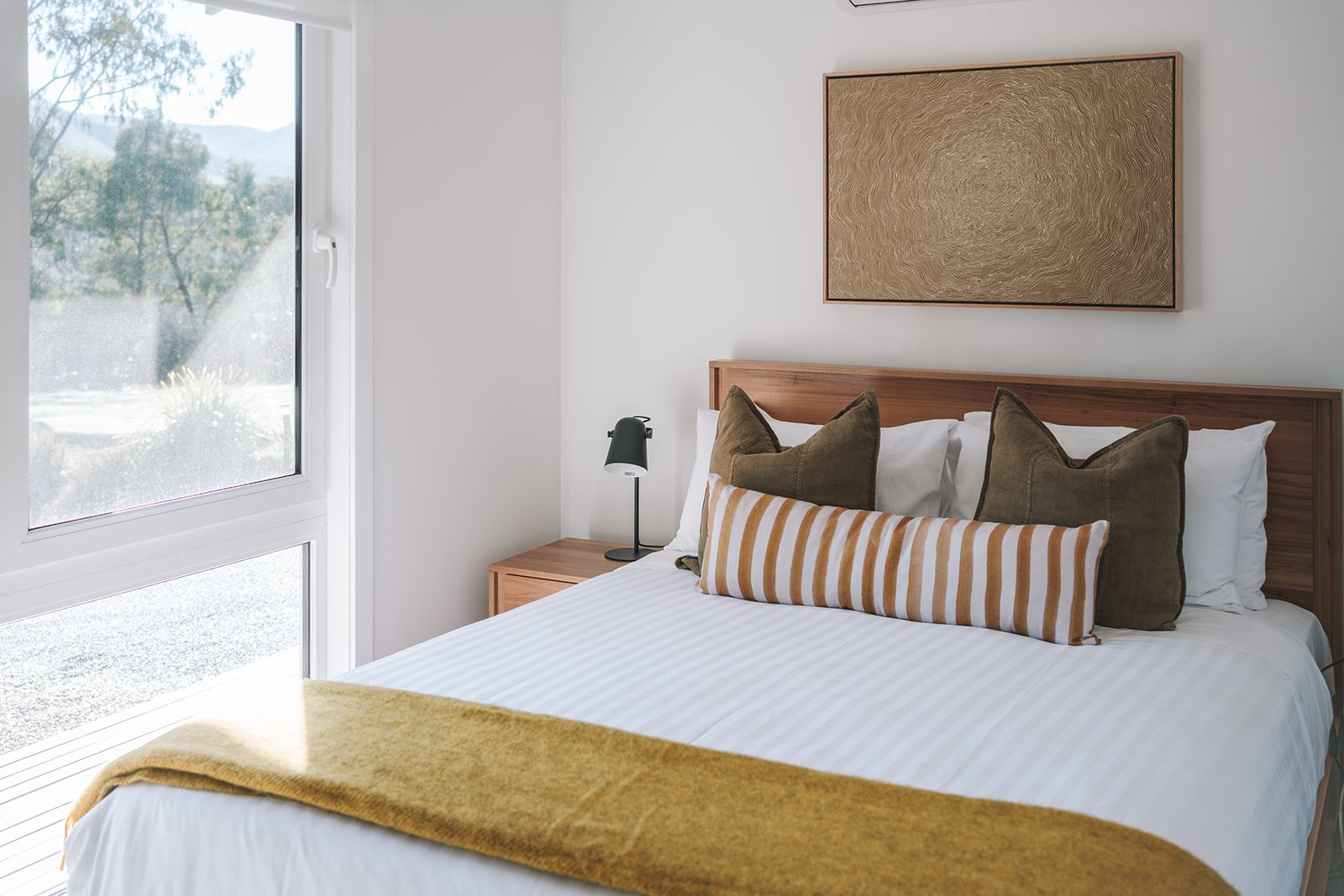 Guest bedroom at Alpine Gum featuring warm timber furniture, olive green cushions, ochre textiles and contemporary Australian artwork.
