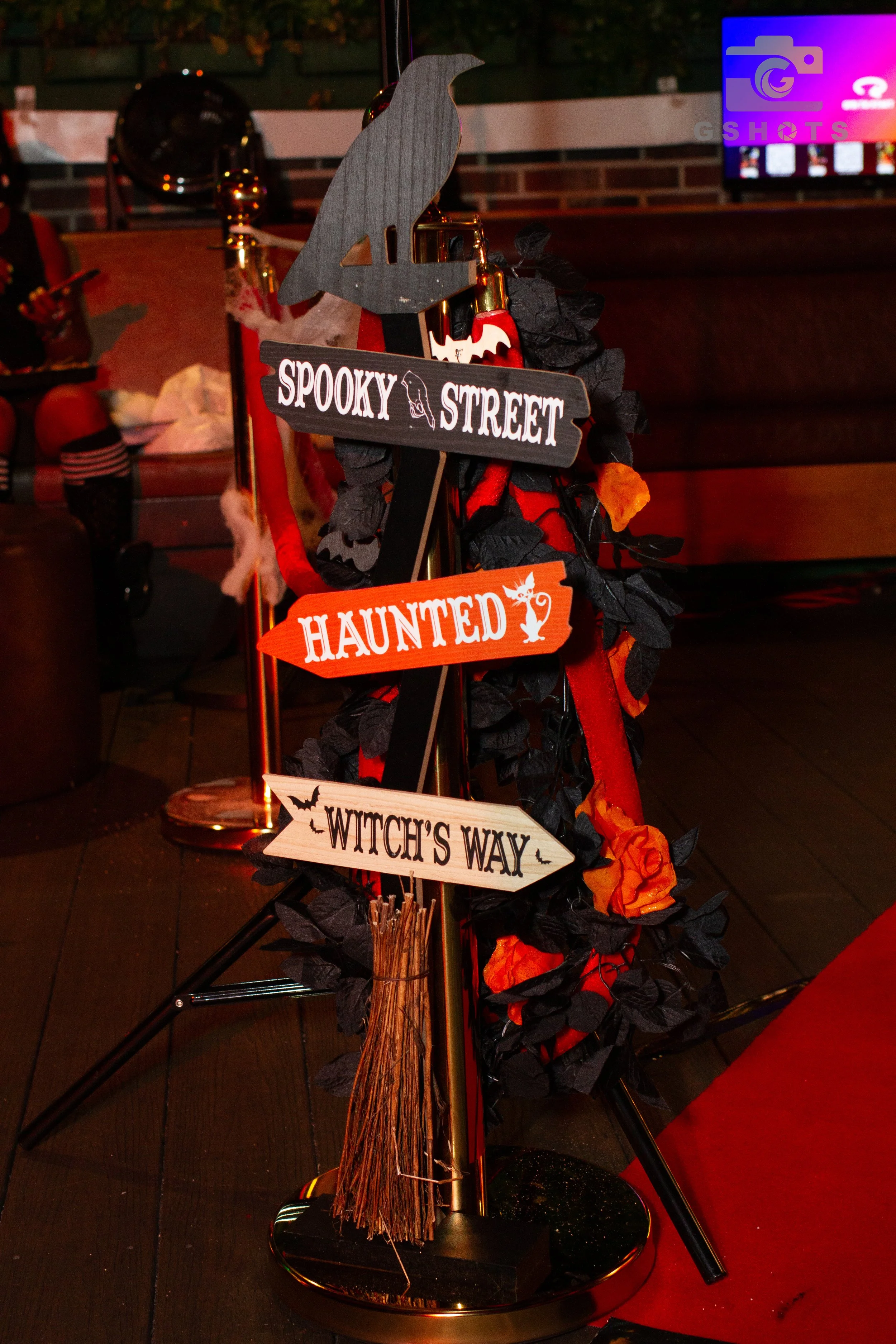 A Halloween-themed signpost with arrows pointing to 'Spooky Street,' 'Haunted,' and 'Witch's Way,' decorated with black and orange leaves and a small broomstick, in a dimly lit indoor setting.