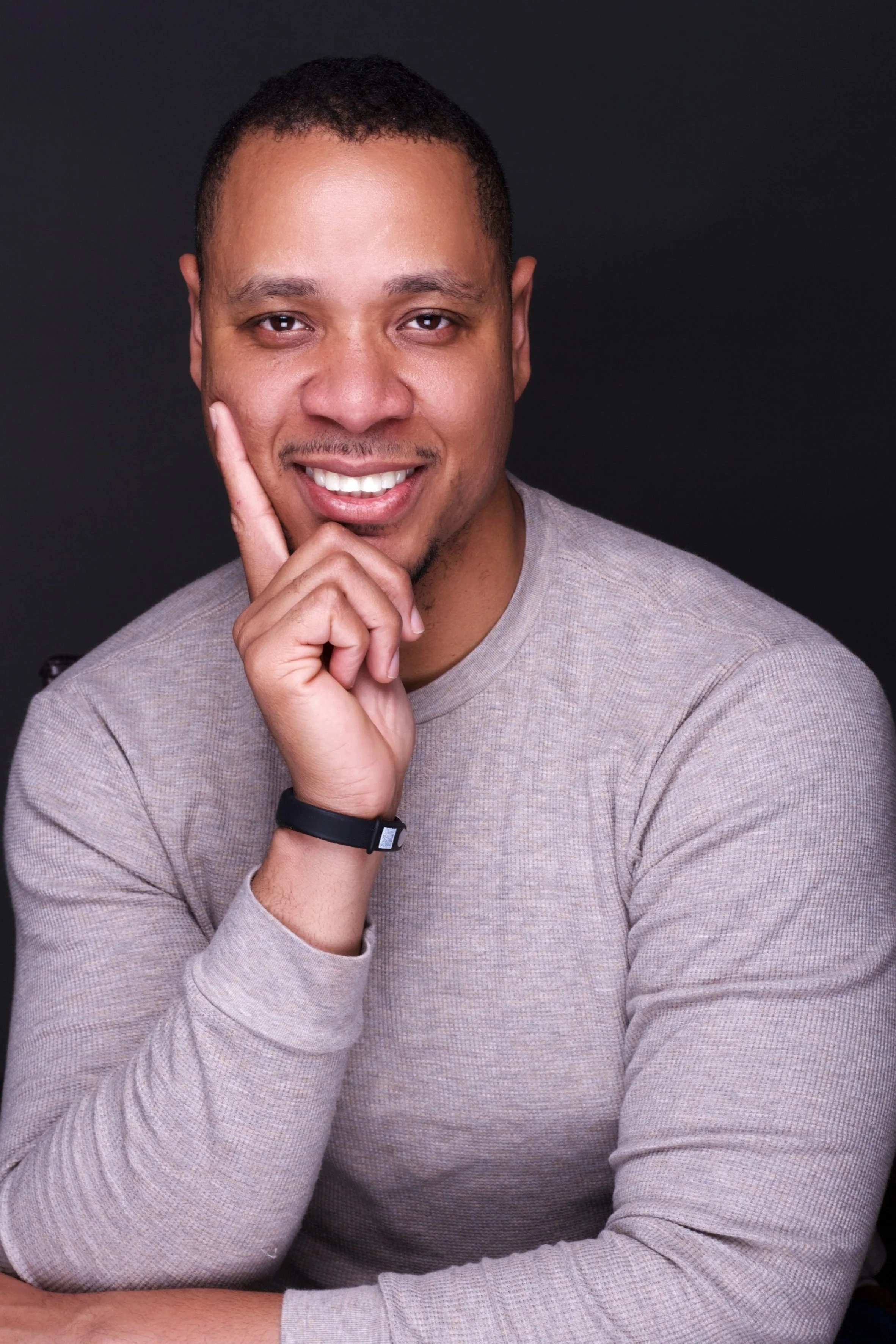A smiling man with short hair, hand resting on face, wearing a beige long-sleeve shirt and a black watch, sitting against a black background.