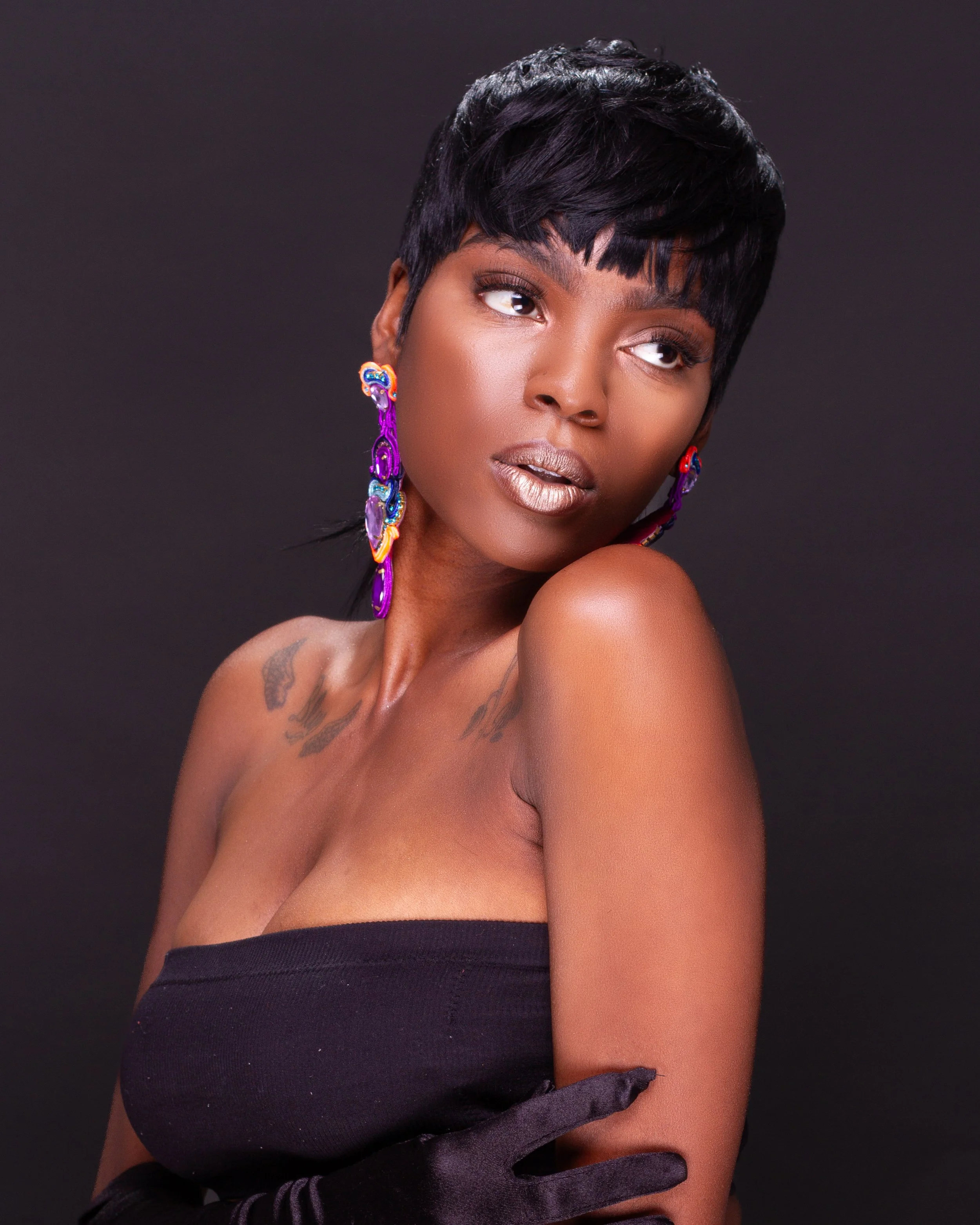 A woman with short black hair and colorful earrings poses against a dark background, wearing a strapless black top and black gloves.
