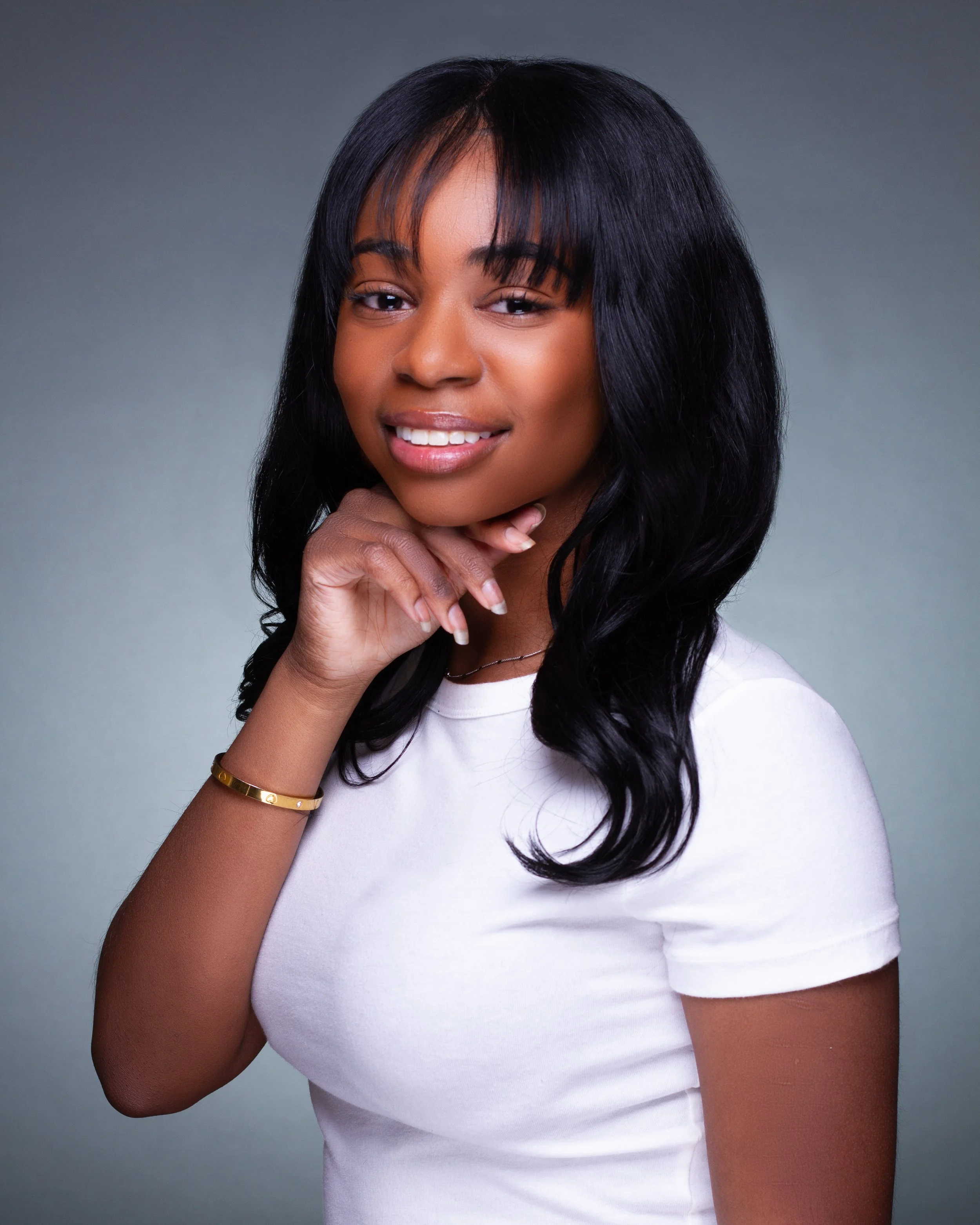 A portrait of a young African American woman with long black hair, smiling and resting her chin on her hand, wearing a white t-shirt and gold bracelet, against a gray background.