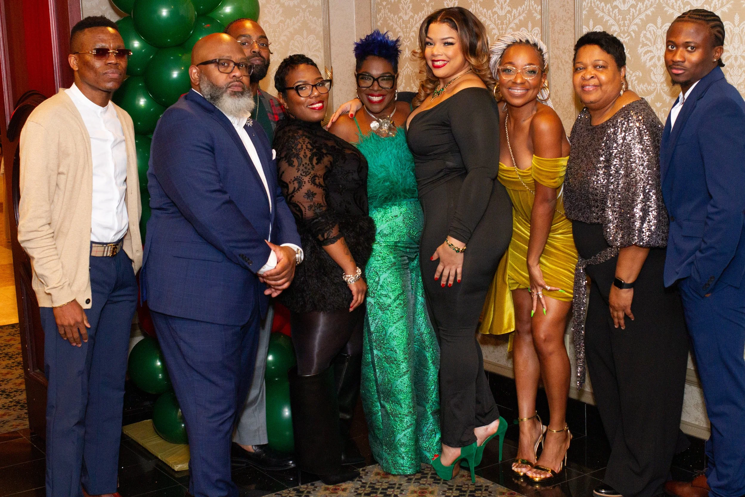 A group of ten diverse people dressed in formal and fashionable attire, standing together in front of a decorative background with green balloons and patterned wallpaper, posing for a photo.