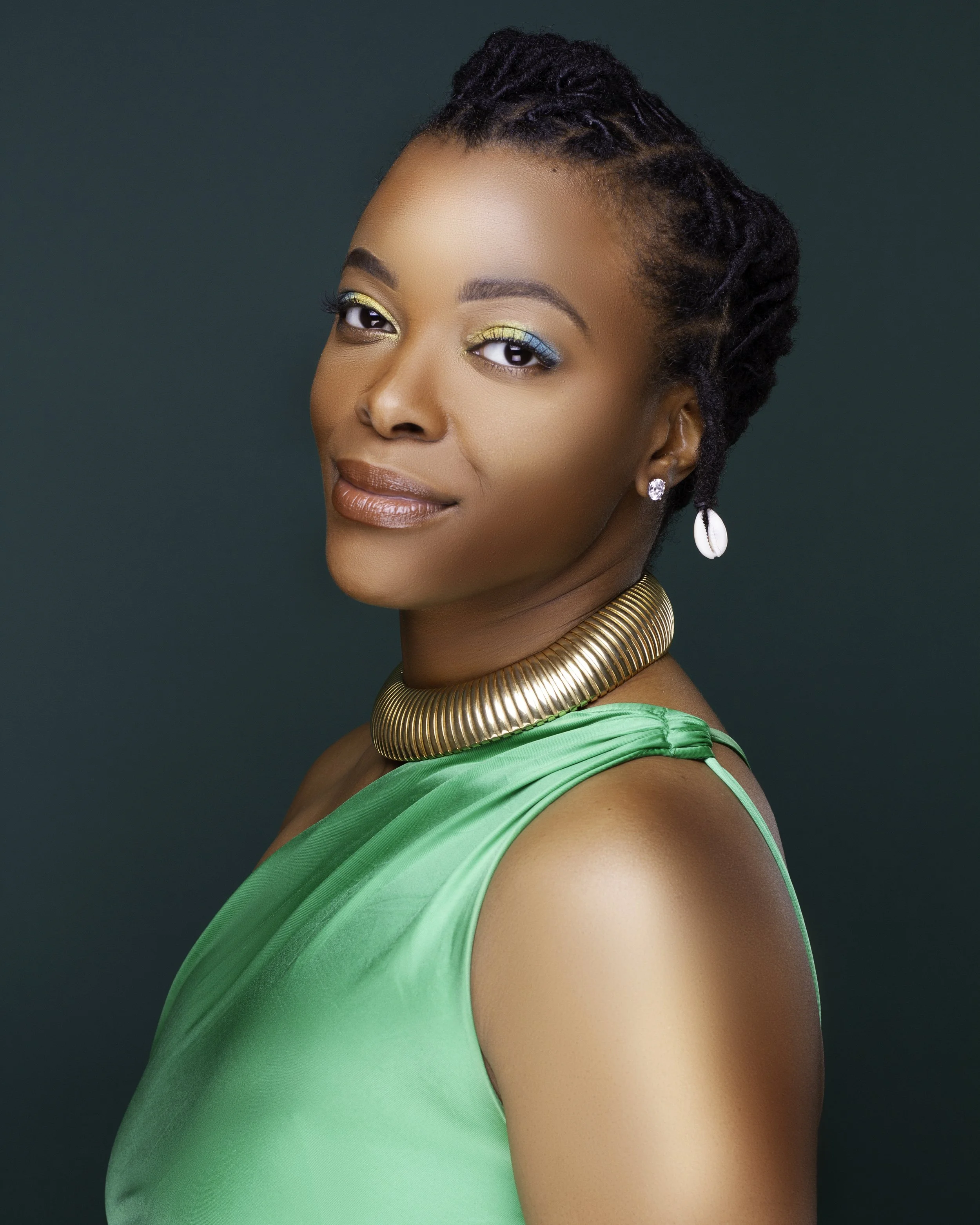 A woman with styled dreadlocks wearing a green dress, gold choker, and shell earrings, posing against a dark green background.