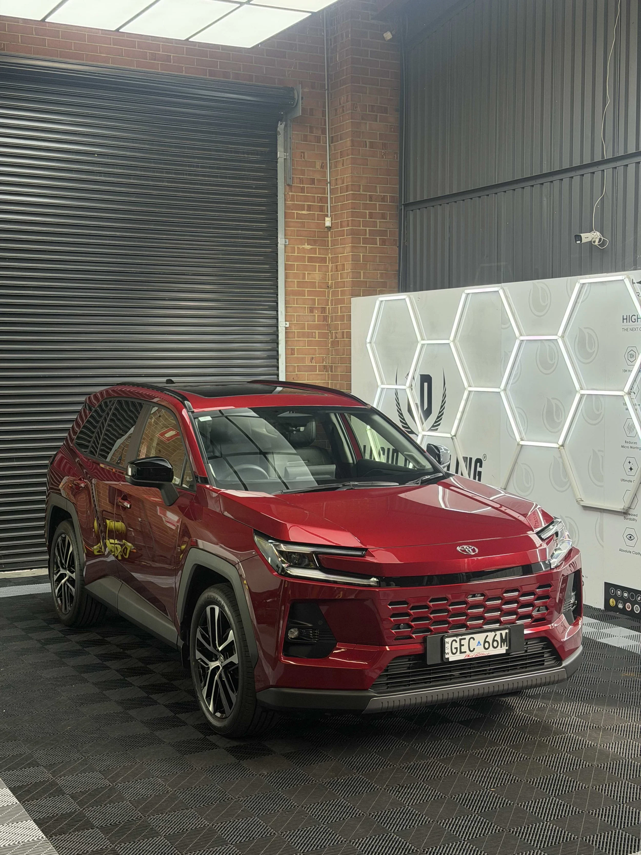 Red Toyota SUV parked indoors in front of a black rolling door and gray brick wall, with a white hexagonal-patterned background display on the right.