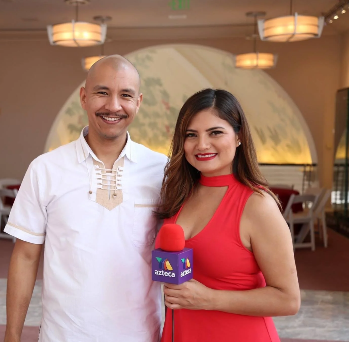 A woman in a red dress holding a microphone with 'azteca' logo, standing next to a man in a white shirt, both smiling in an indoor setting with warm lighting and decorative ceiling fixtures.