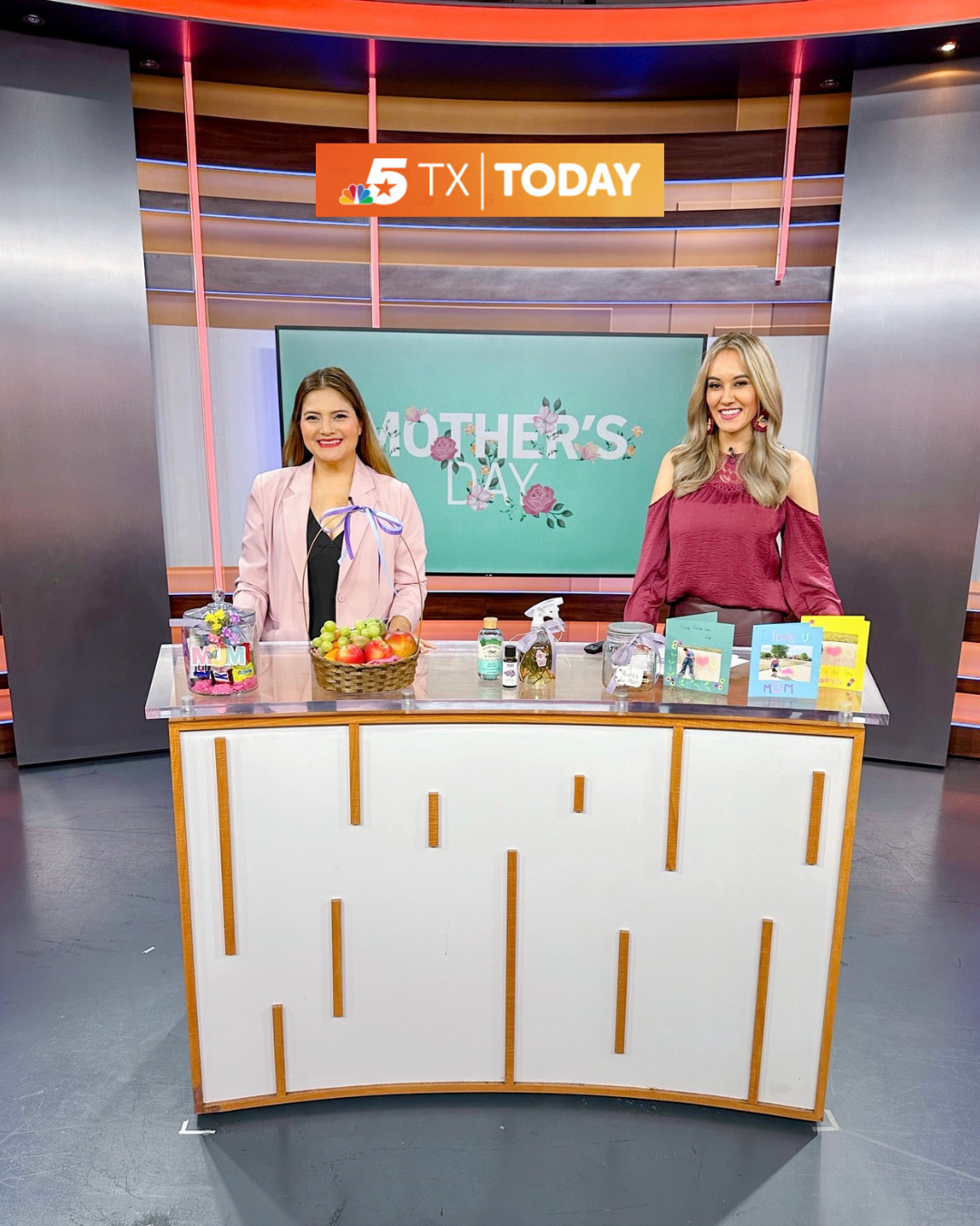 Two women are standing behind a white news desk decorated with fruits, flowers, and greeting cards, on a television set celebrating Mother's Day. A large screen behind them displays the words 'Mother's Day' with floral graphics, and a sign above shows '5 TX TODAY'.