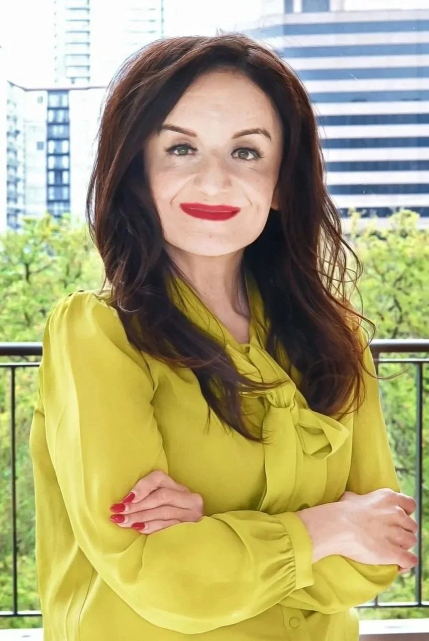 A woman with long brown hair and red lipstick, wearing a yellow blouse, standing outdoors on a balcony with a cityscape and green trees in the background.