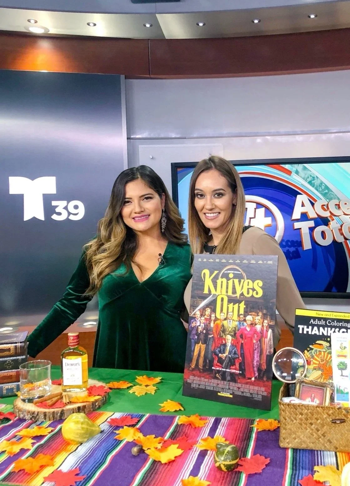 Two women standing behind a table with fall decorations, a sign for the movie 'Knives Out,' and various items, in a TV studio setting.