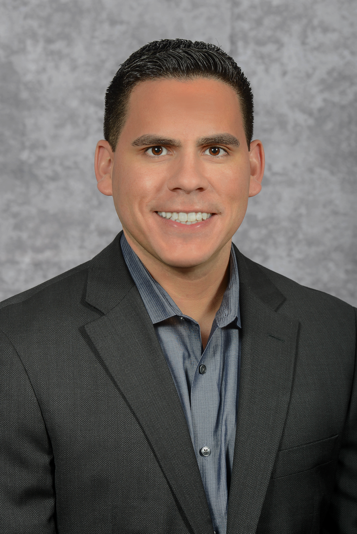 Professional headshot of a smiling man in a dark suit jacket and blue dress shirt, gray background.