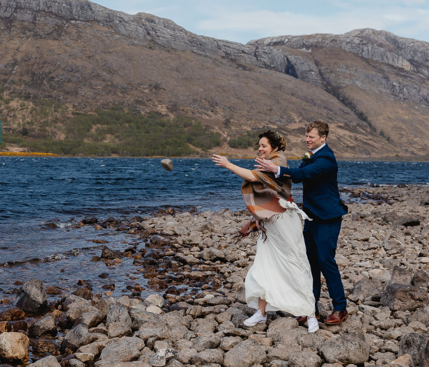 A bride and groom throwing a stone into a lake on a rocky shoreline, with mountains in the background.