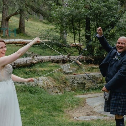 A bride and groom dressed in a wedding dress and kilt, holding a traditional Scottish sword, are playfully pulling a rope in a wooded outdoor setting during their wedding celebration.