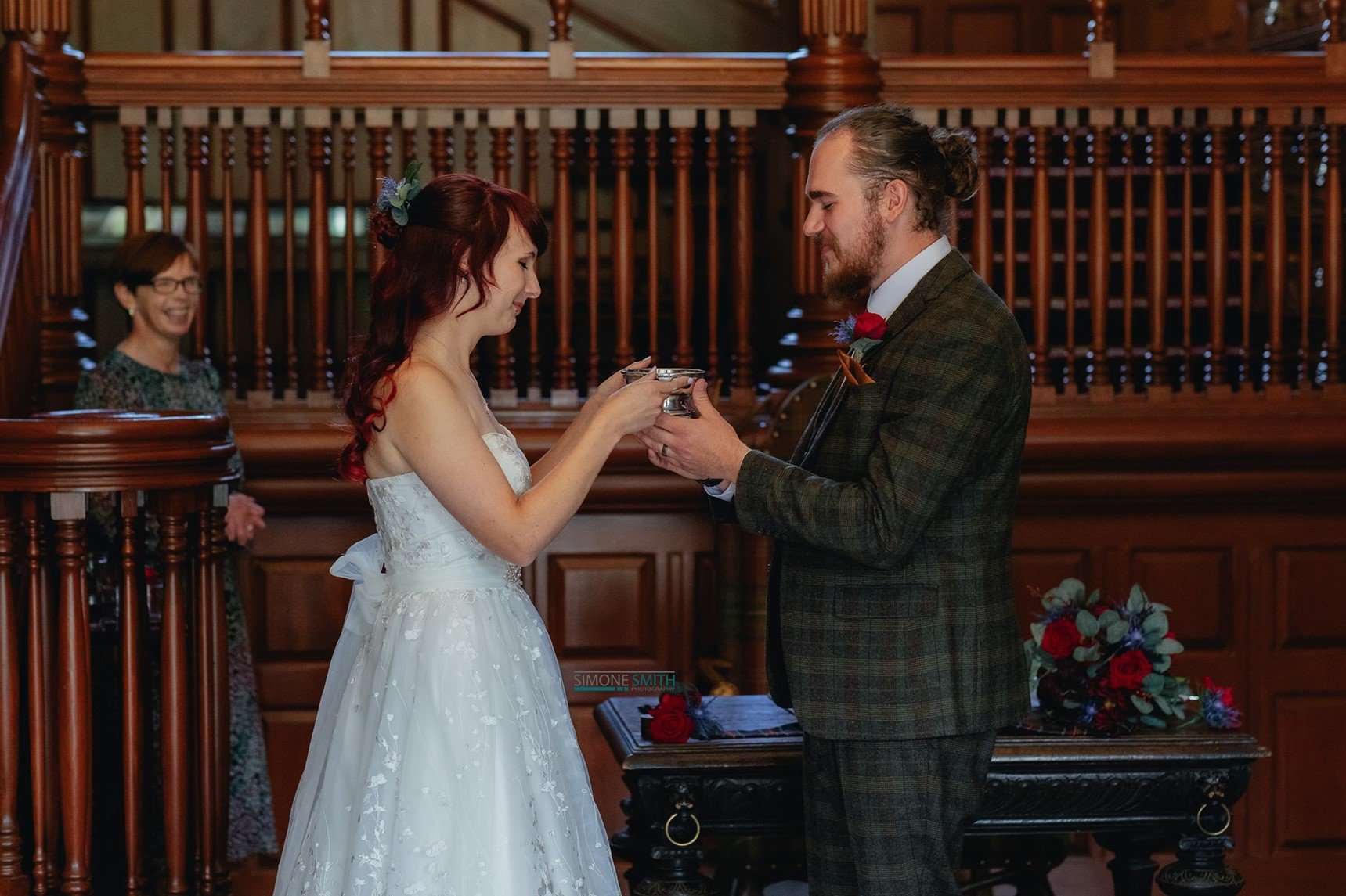 A couple exchanging wedding vows in a ceremony, with a woman in a white dress and a man in a suit, both smiling, while a woman in the background observes.