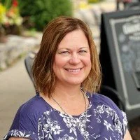 Smiling person with short hair wearing a blue floral top, sitting outdoors.