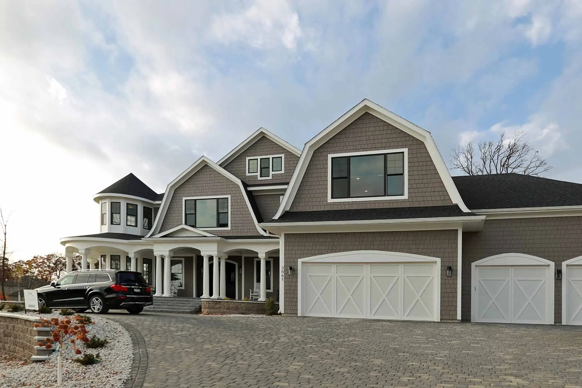 A large, multi-story house with gray siding, white trim, and a black roof. The house features several gables, multiple windows, a porch with white columns, and a white double garage door. A black car is parked in the driveway, and the yard has decora