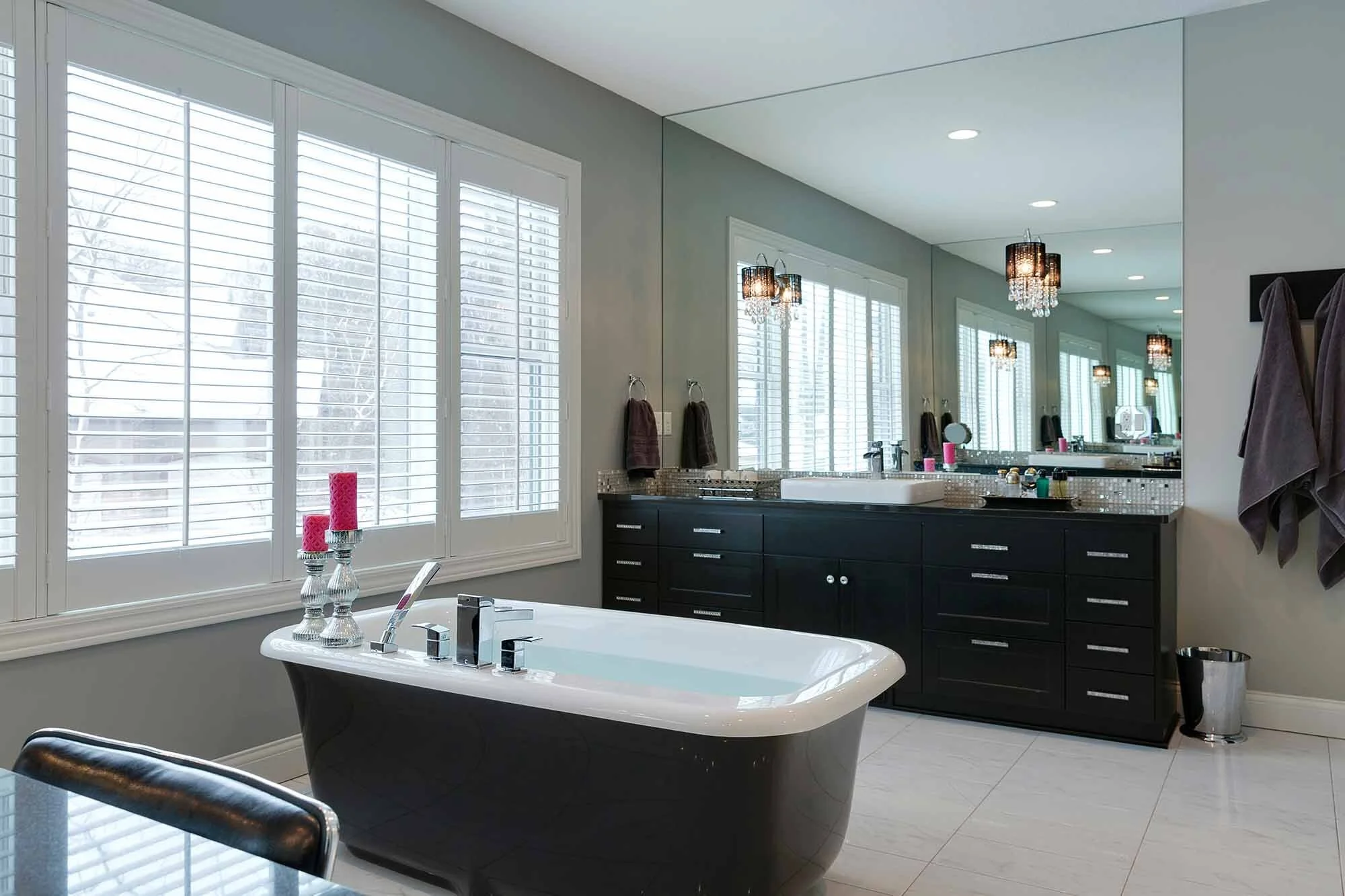 Modern bathroom with black vanity, large mirror, white bathtub, and window with white shutters.