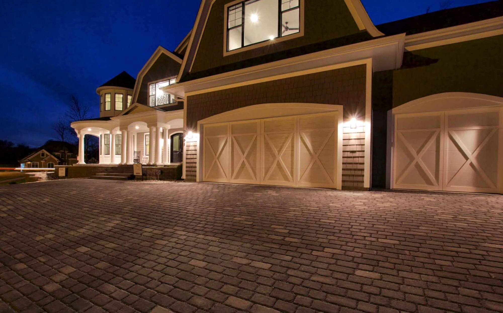 Night view of a large, elegant house with a brick driveway, illuminated windows, and exterior lights highlighting the porch and garage doors.