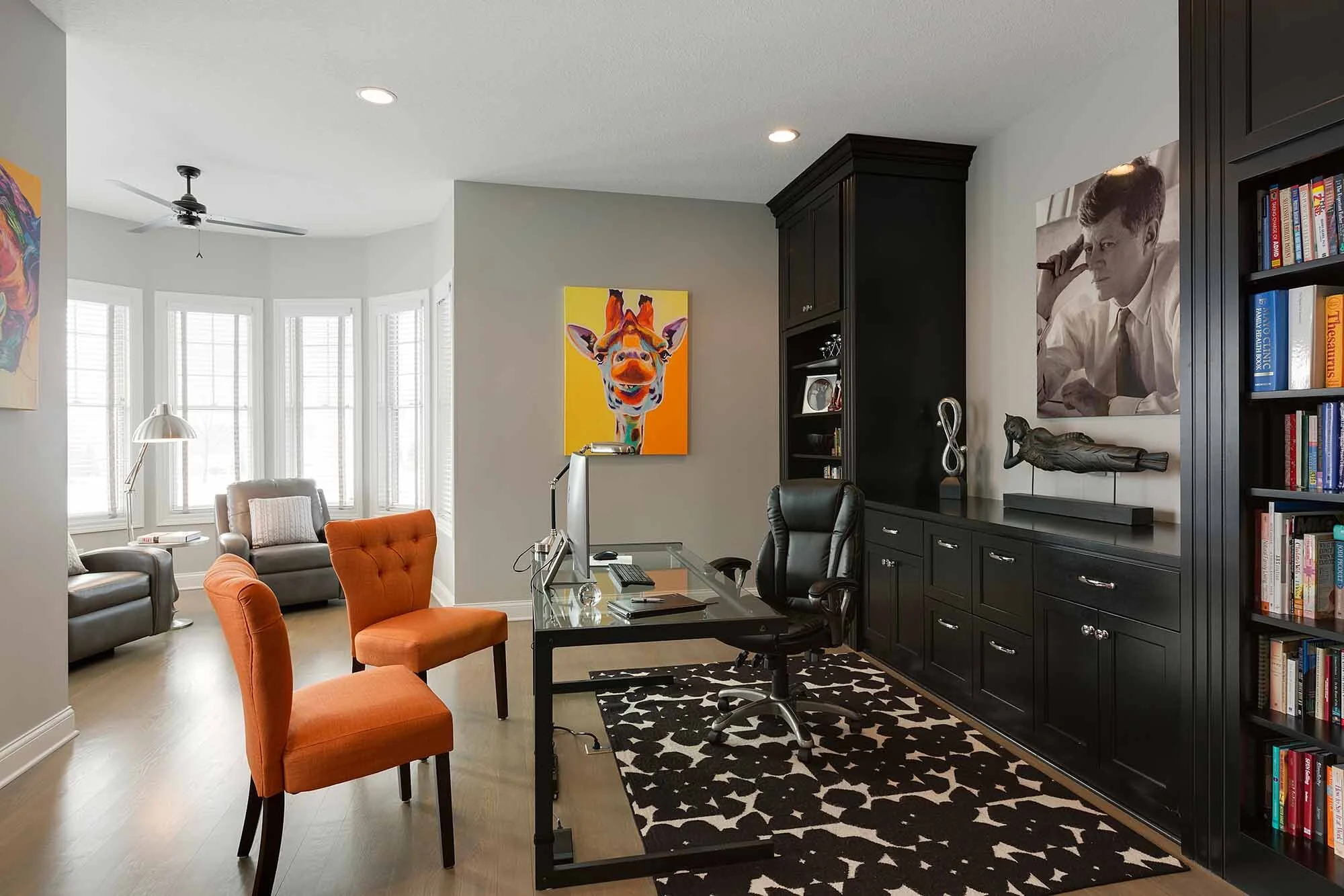 Home office with a black desk, black office chair, orange chairs, a black rug with white patterns, built-in black shelving unit with books and sculptures, colorful animal painting on the wall, black and white framed photograph, and a reading nook wit