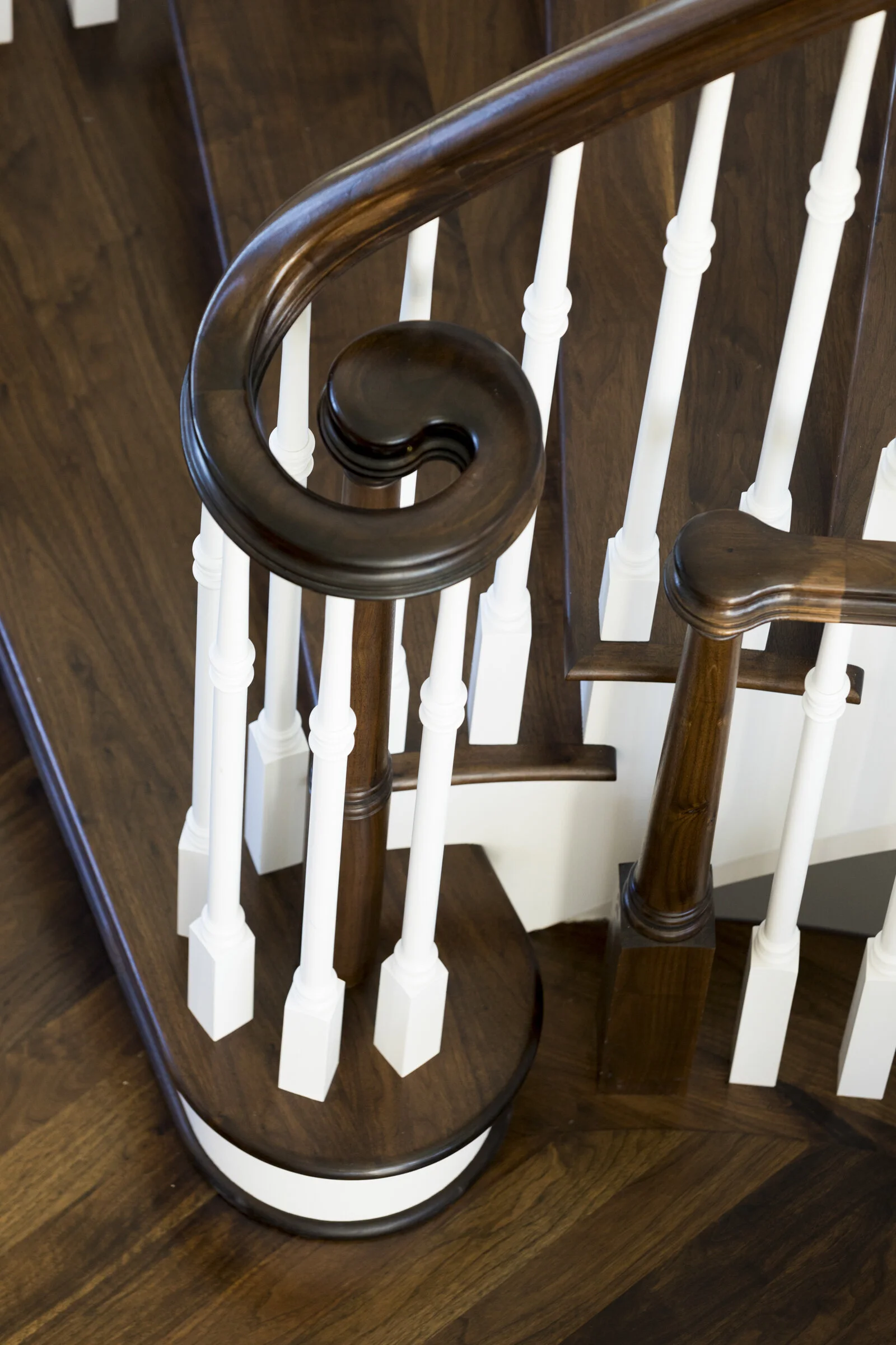 Top view of a wooden staircase handrail with a spiral design on a dark wood staircase with white balusters.