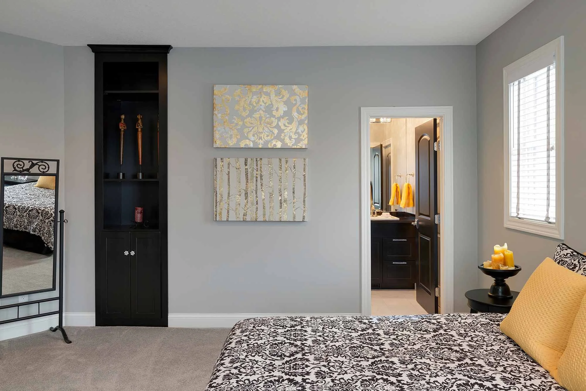 Bedroom with black and white patterned bedspread, yellow pillows, small black side table with candles, gray wall with two canvas paintings, mirror, open door to bathroom with dark vanity and yellow towels, window with blinds.
