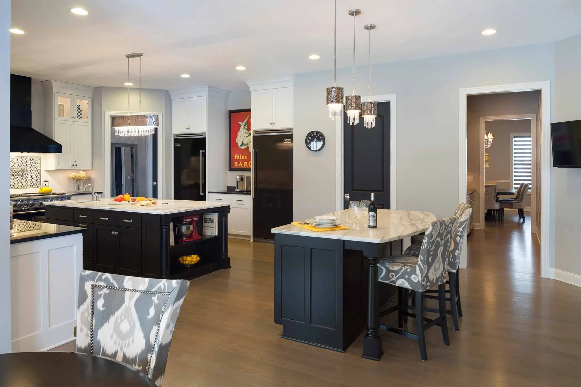 Modern kitchen with black and white cabinets, a large island with fruit, and a small marble-topped eating area with patterned chairs.