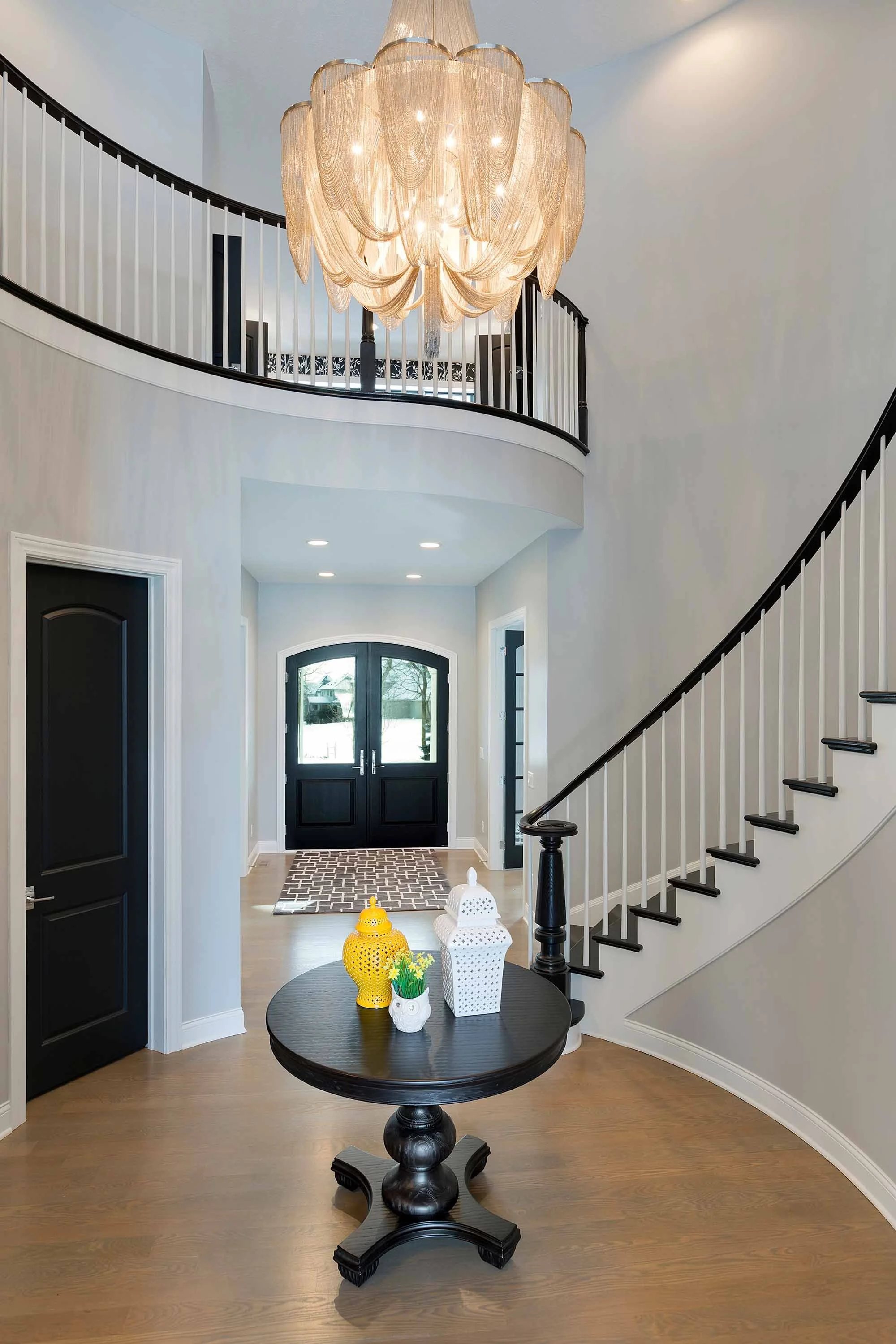 Elegant foyer with a black double front door, white walls, wooden flooring, a round black table with decorative vases and lanterns, and a large chandelier hanging from the ceiling.