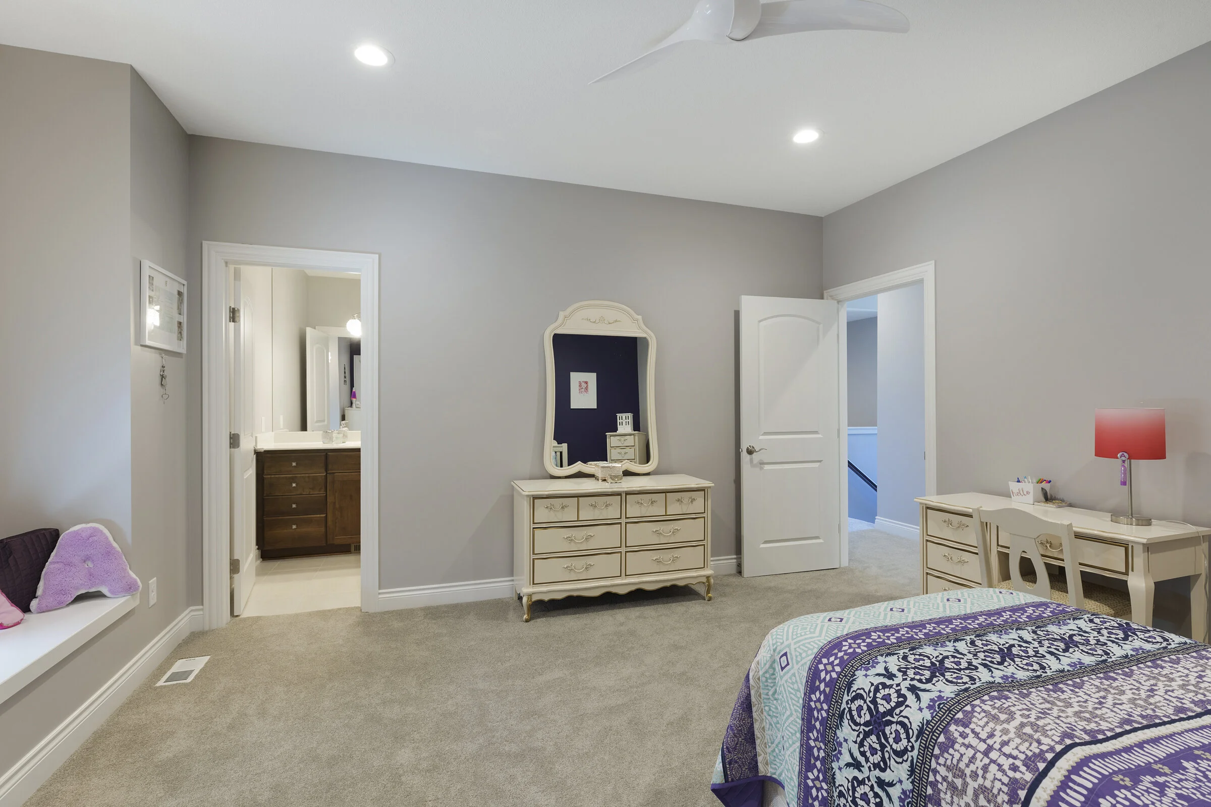 A bedroom with a beige carpet, a bed with a purple, teal, and white quilt, a white desk with a pink lamp, a white dresser with a mirror, and an open door showing a bathroom. The walls are painted light gray.