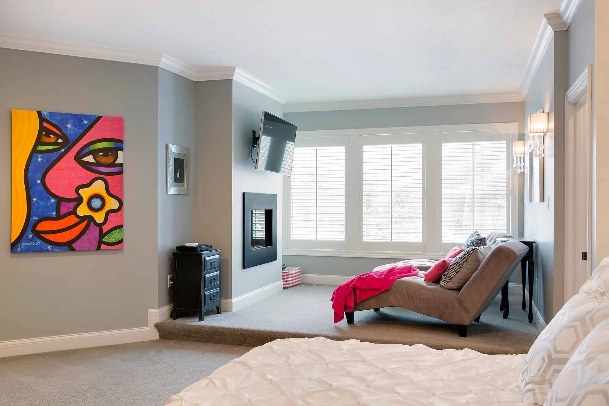 Living room with grey walls, large window with white shutters, beige sofa with pink and brown pillows, wall-mounted TV, colorful abstract face painting on the wall, and a fireplace or wall feature beneath the TV.