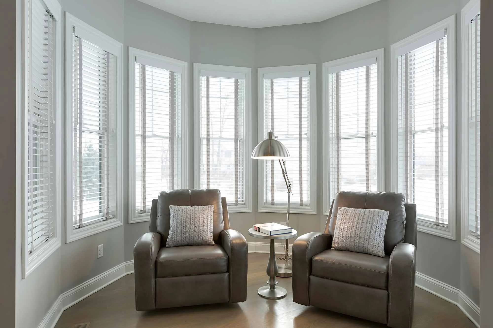 A cozy reading nook with two brown leather armchairs, a small round side table with books, and a tall floor lamp, surrounded by multiple large windows with white blinds.