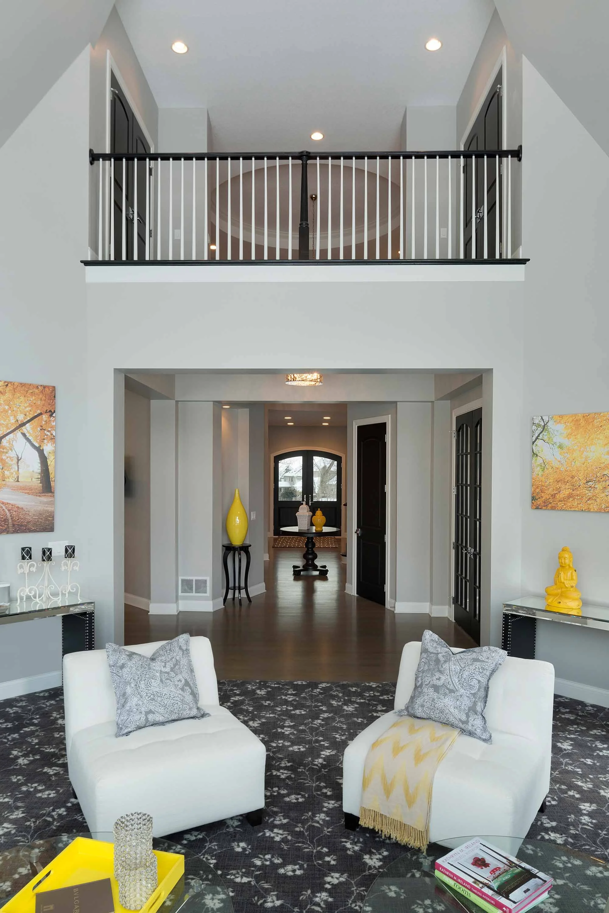 Living room with two white armchairs, yellow and gray decorative pillows, dark patterned carpet, and two side tables with yellow and silver decorations. Background features a hallway with hardwood floors, decorated with yellow and white vases, leadin