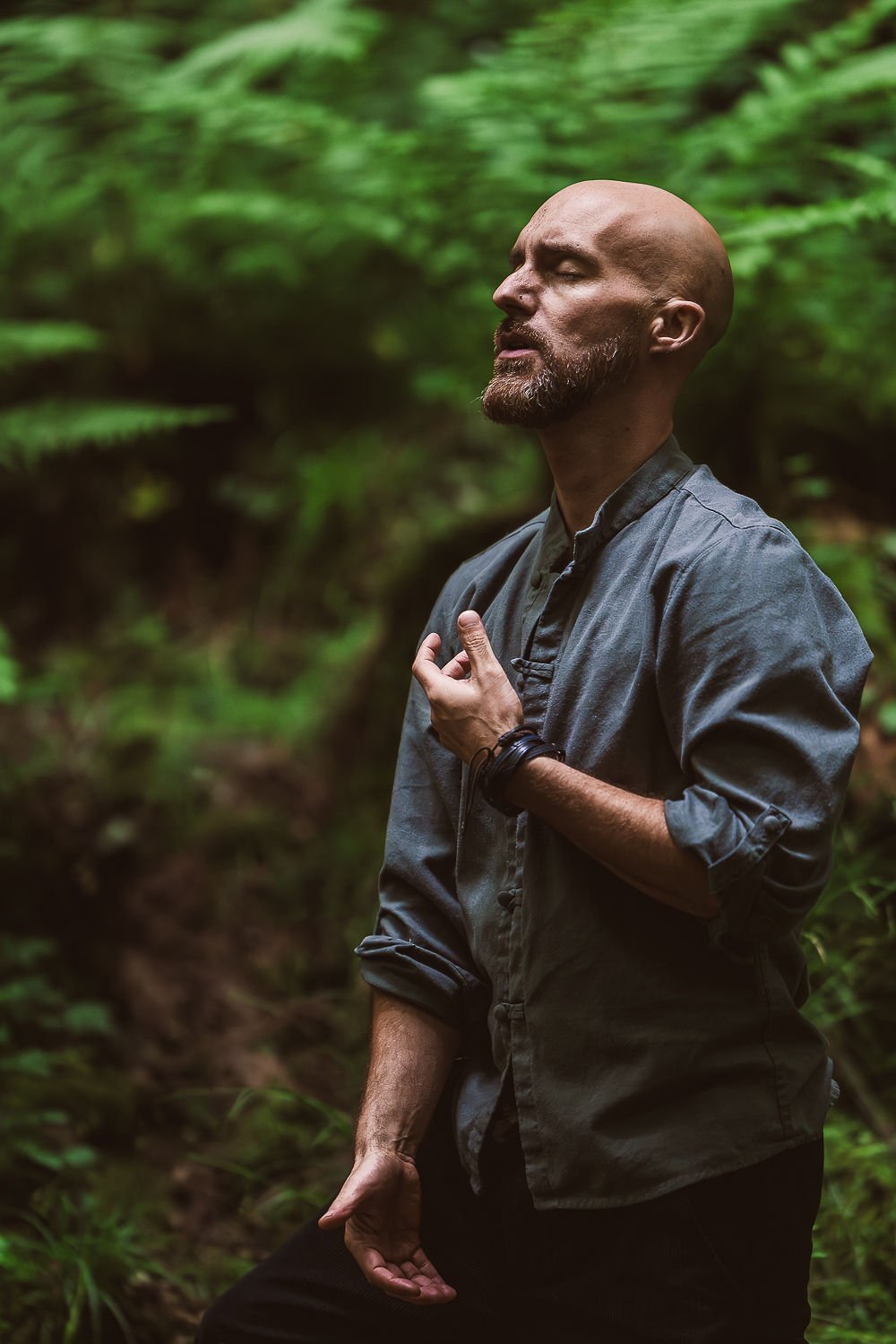A man with a beard and shaved head practicing meditation outdoors in a lush green forest, with one hand on his chest and the other resting down.