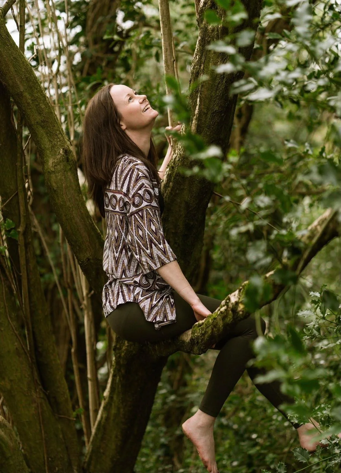 A woman sitting on a tree branch in a forest, looking up and smiling.