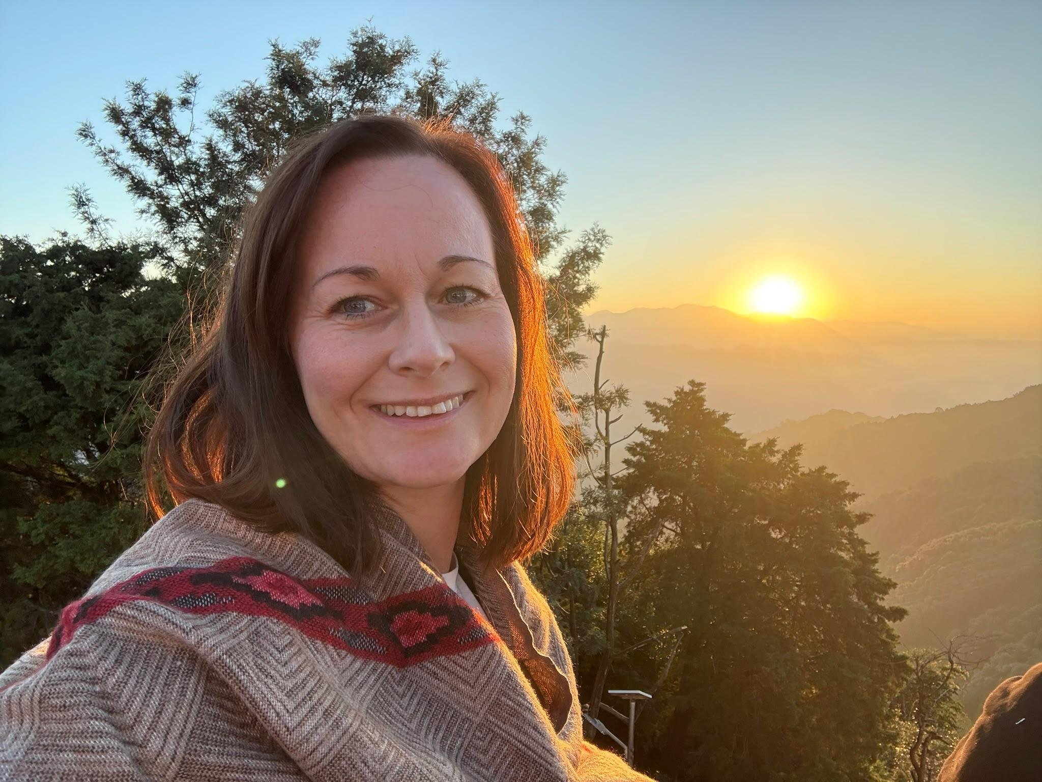 A woman smiling outdoors at sunset with trees and mountains in the background.